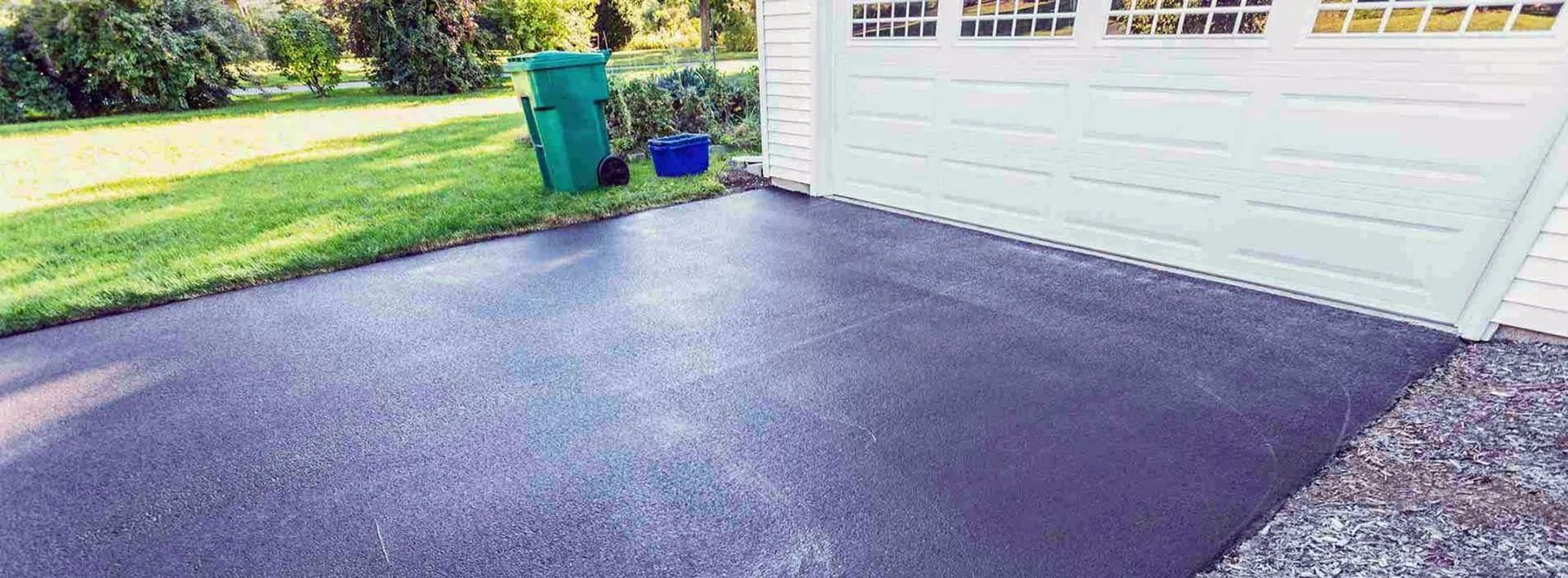 Freshly paved dark driveway beside a white garage, with green trash bins near the lawn.