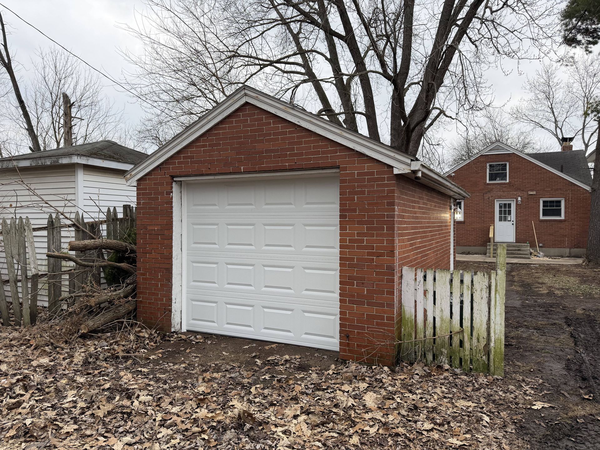 Brick garage with white door and weathered fence; residential setting.