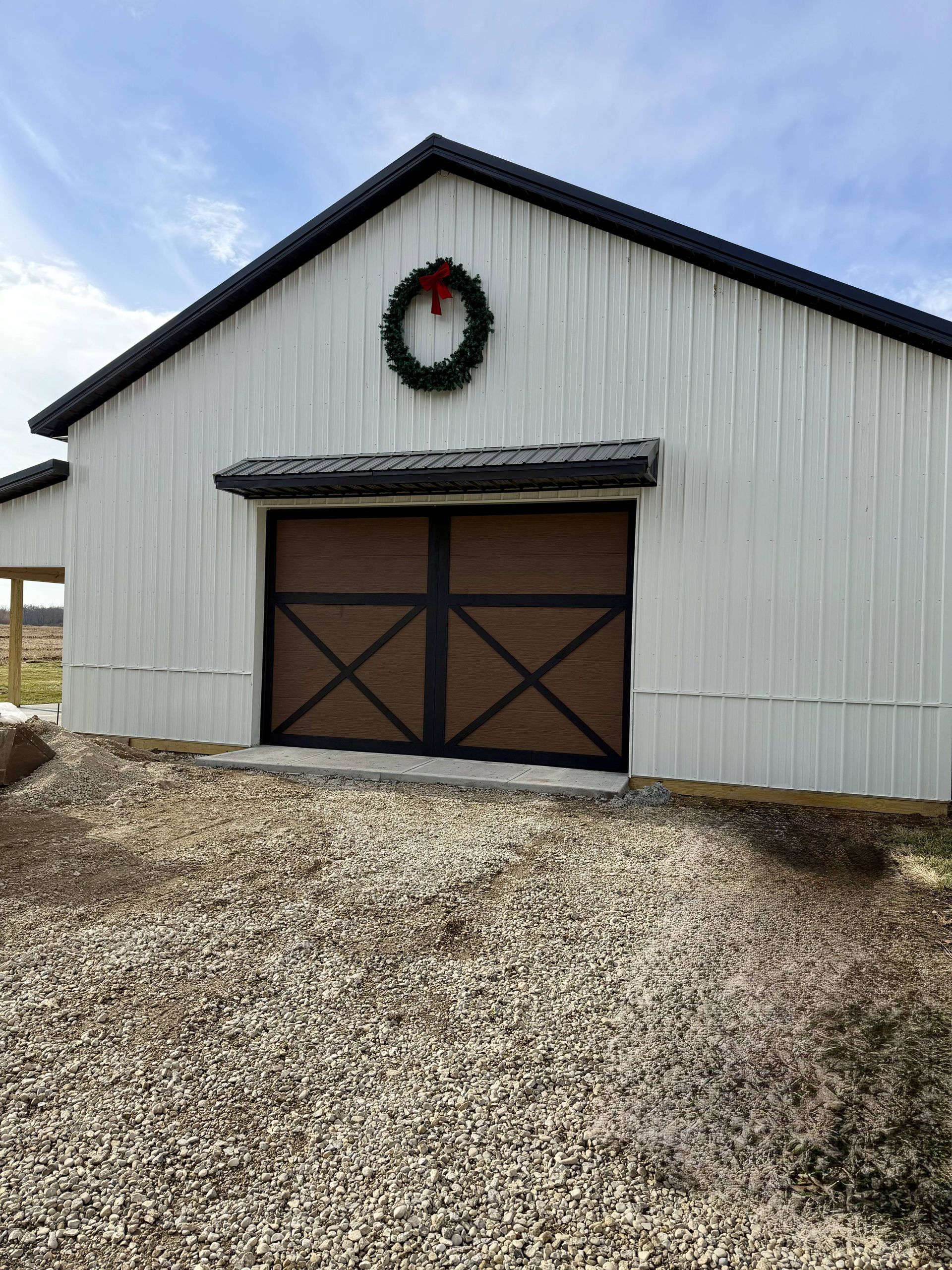White barn with brown door and a Christmas wreath hanging above. Gravel in foreground.