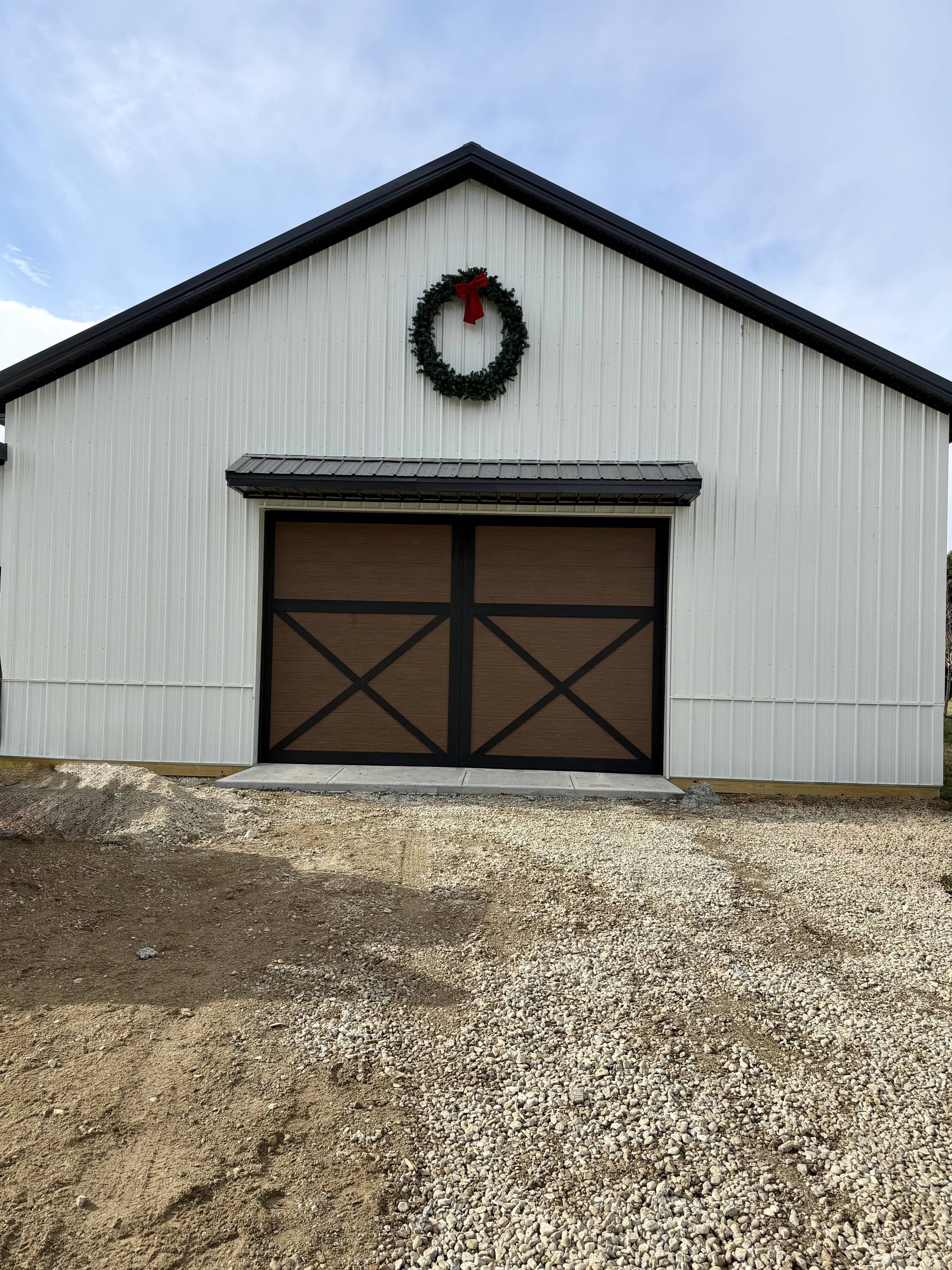 White barn with brown doors, a wreath above, and gravel ground.