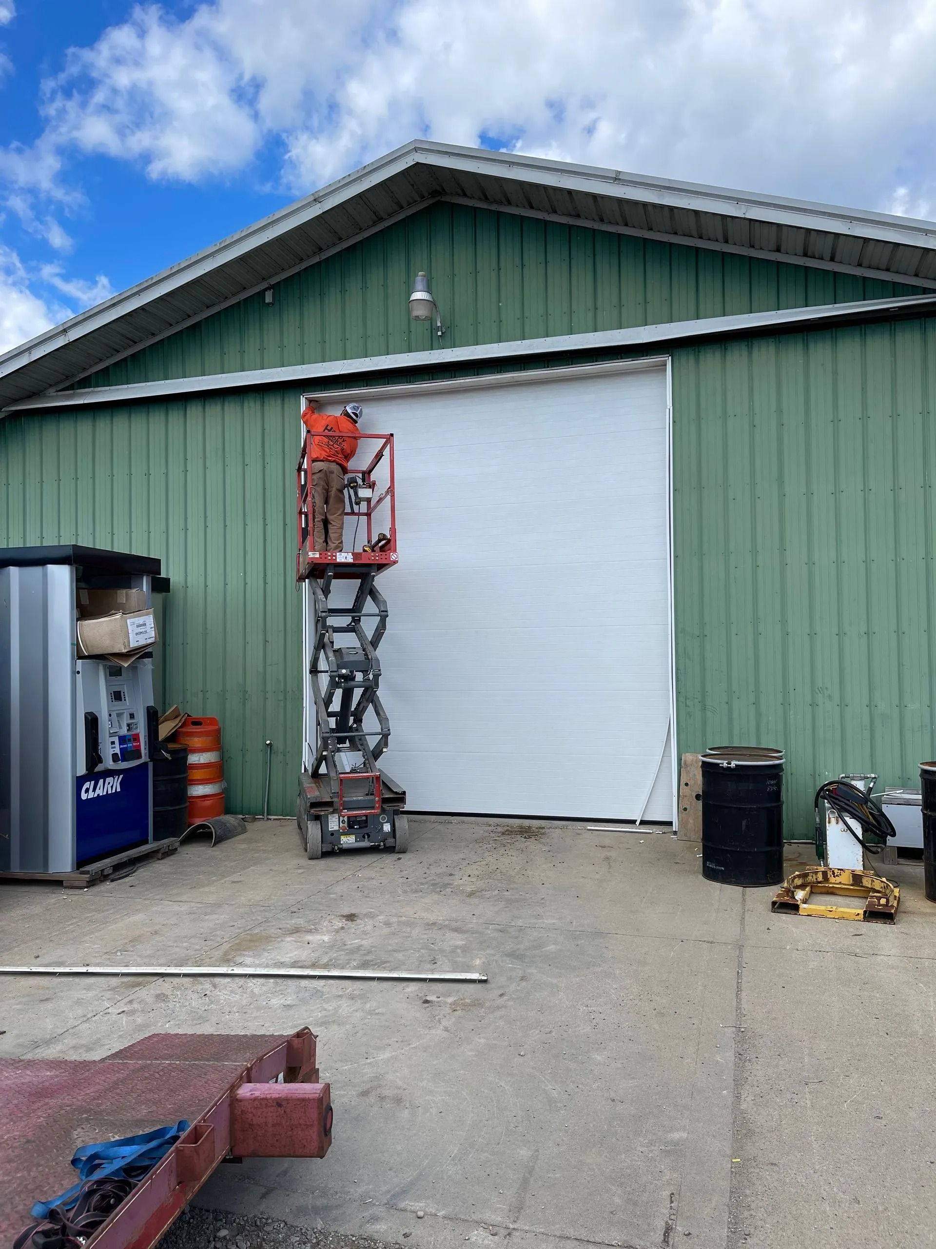 A man in a lift working on a large white door of a green industrial building.