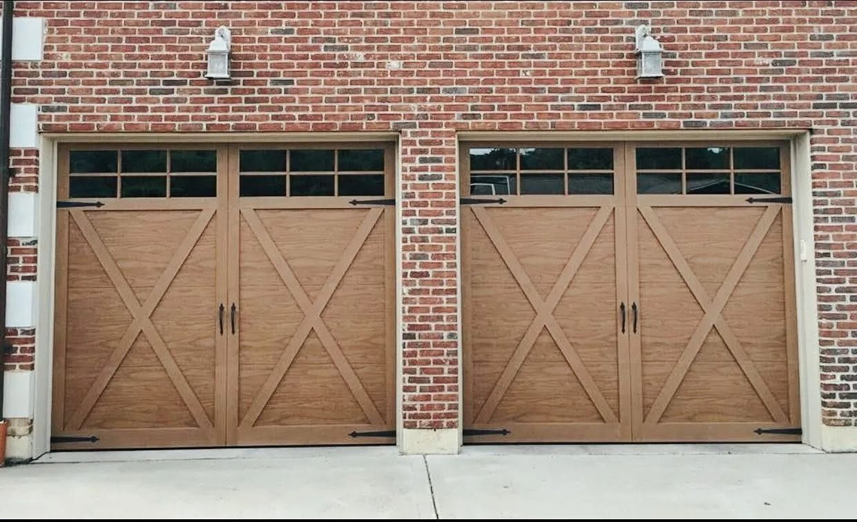 Two tan wooden garage doors with cross design, brick building background, gray concrete.