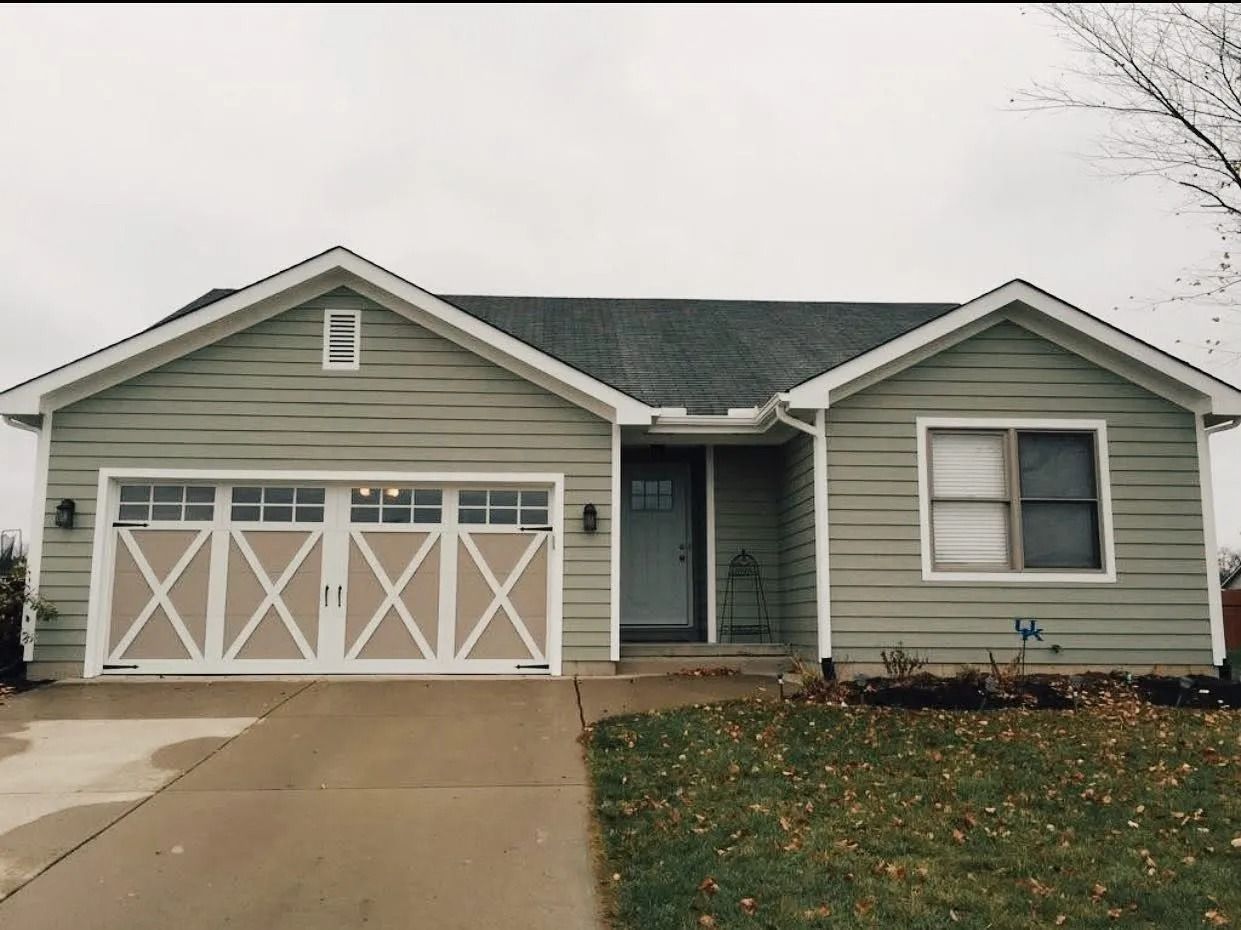 Single-story house with green siding, white trim, and tan garage doors. Gray roof, gray sky, and a concrete driveway.