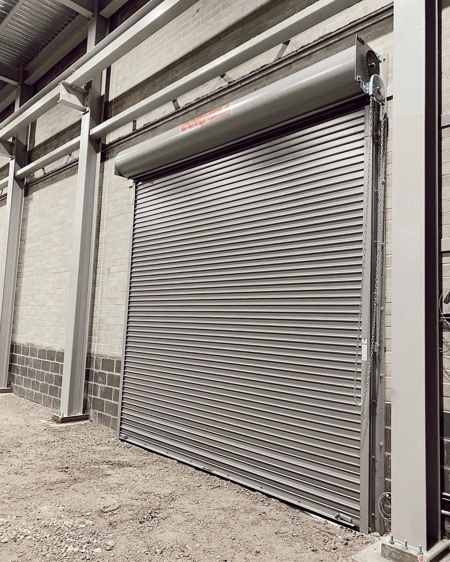 Roll-up metal security door on a brick building with a gravel ground, under a steel structure.