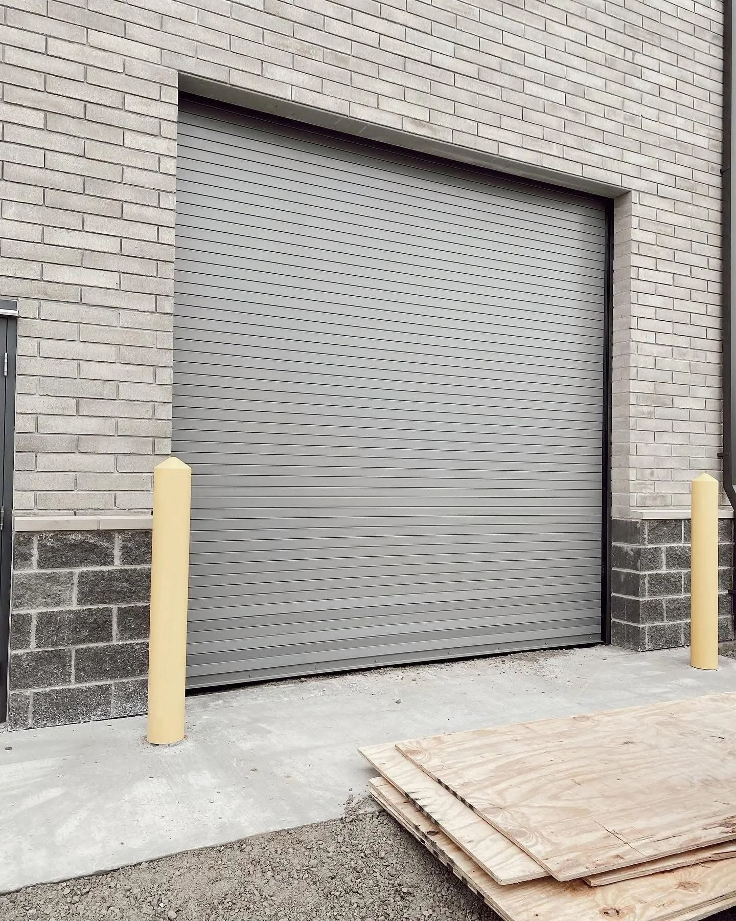 Gray industrial roll-up door in brick building with two yellow bollards. Plywood on ground.