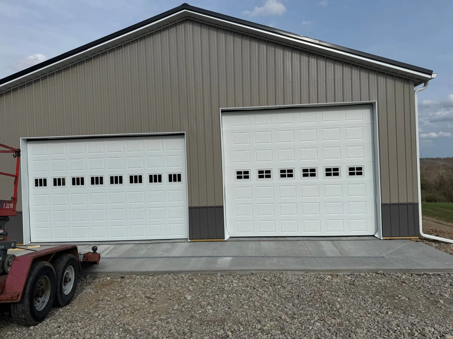 Two white garage doors on a gray metal building with a concrete apron; a trailer is parked nearby.