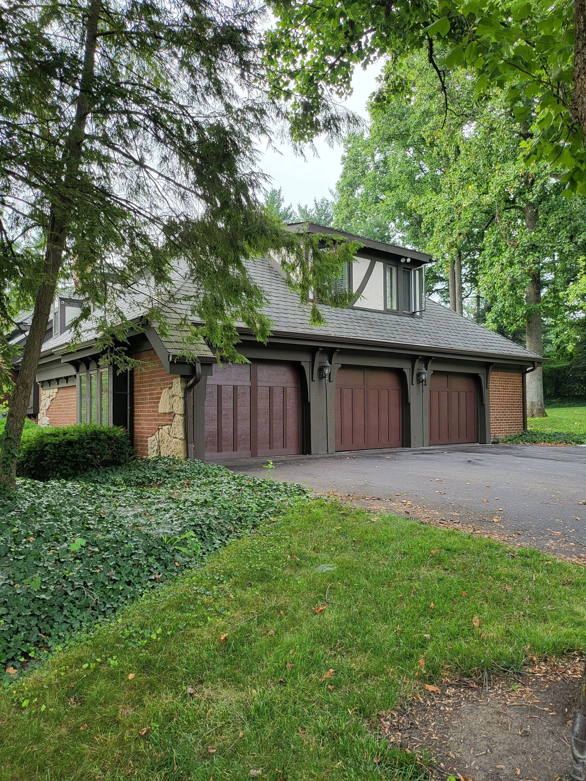 Three-car garage with brown doors, brick siding, and a gabled roof. Green trees and ivy surround the building.