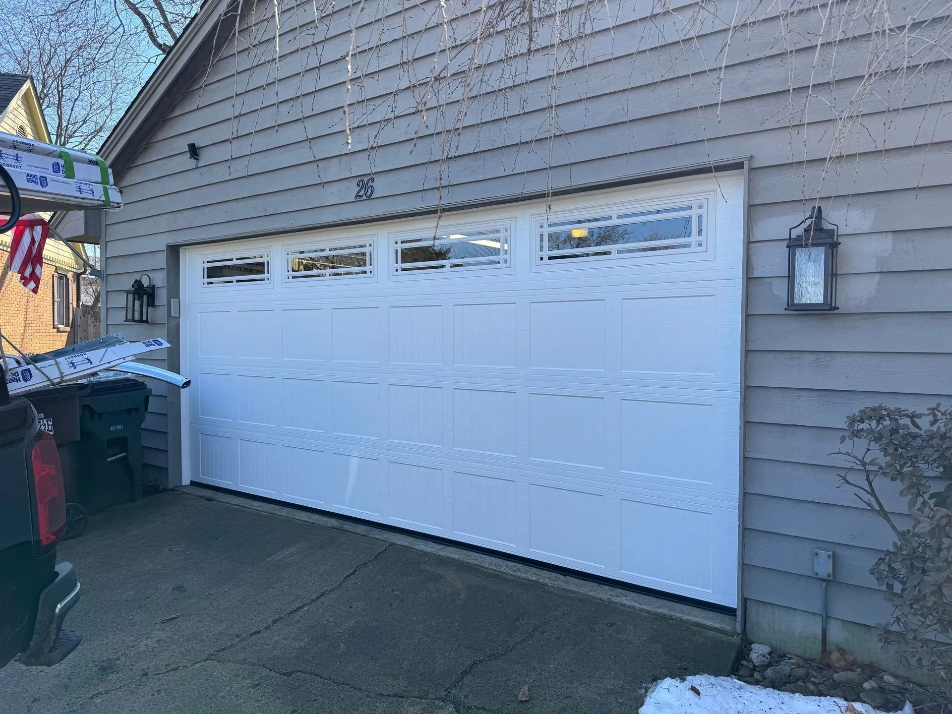 White garage door with rectangular windows, mounted on a gray house. Two lanterns are mounted on the wall.