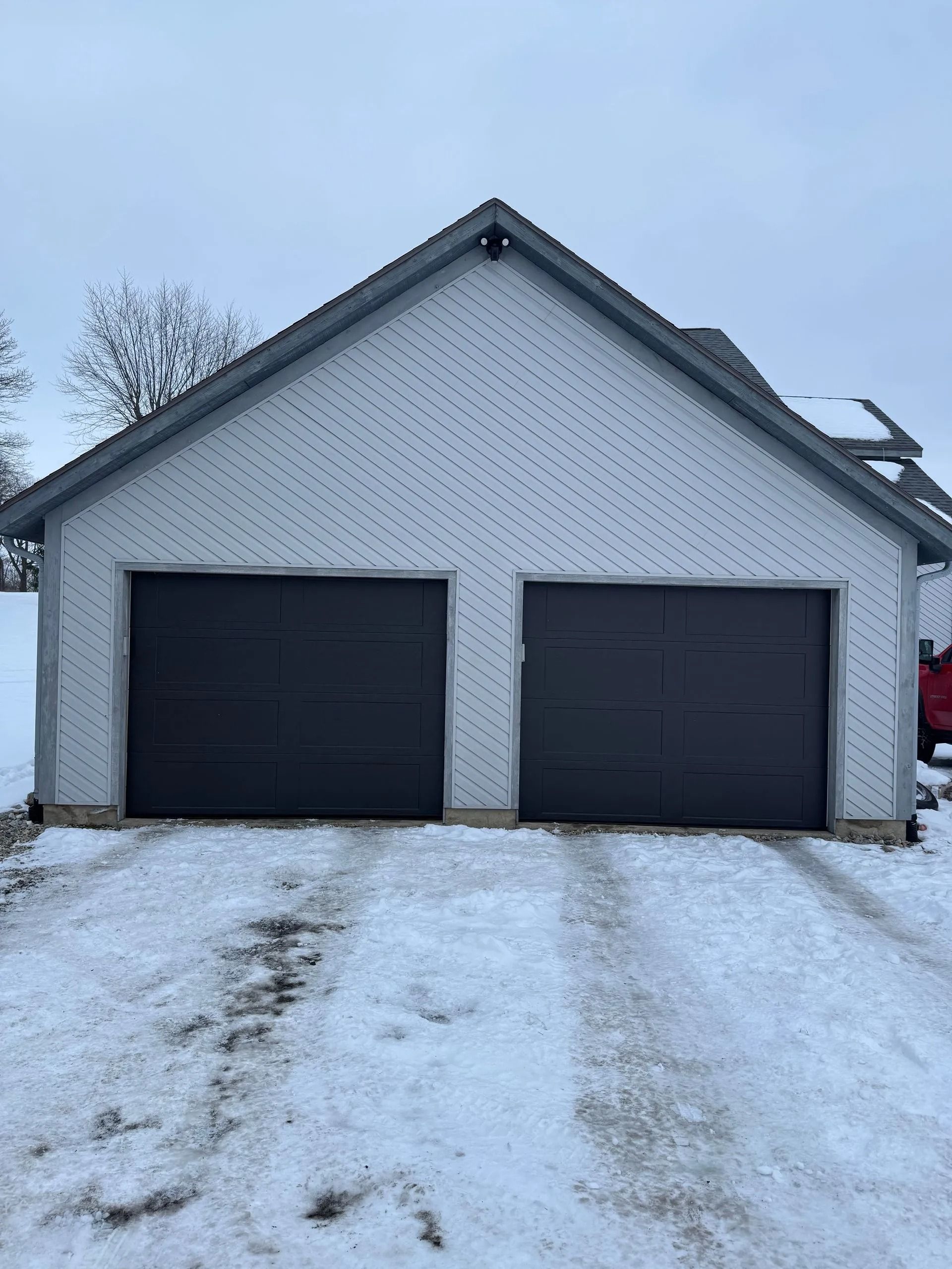 Gray two-car garage with dark gray doors, a white roof, and a snowy driveway.