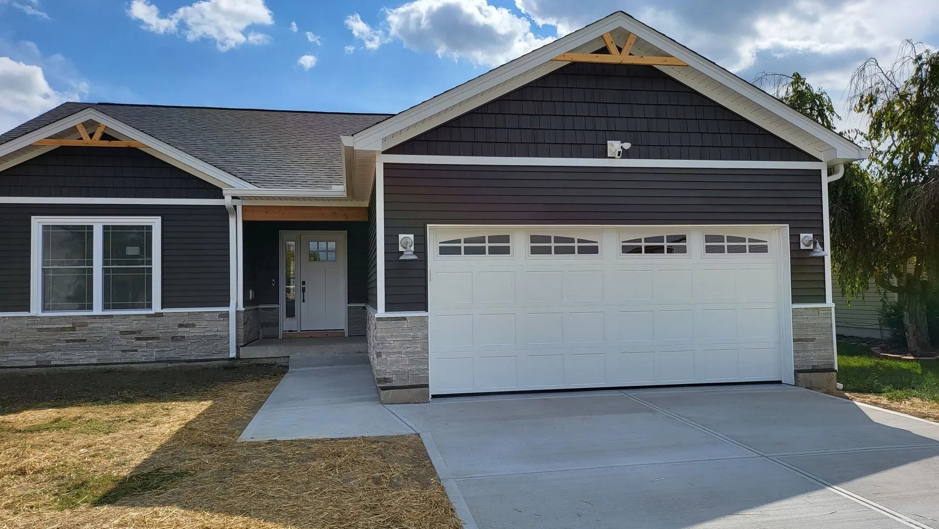 Single-story house with dark brown siding, white garage door, and light stone accents, under a cloudy sky.