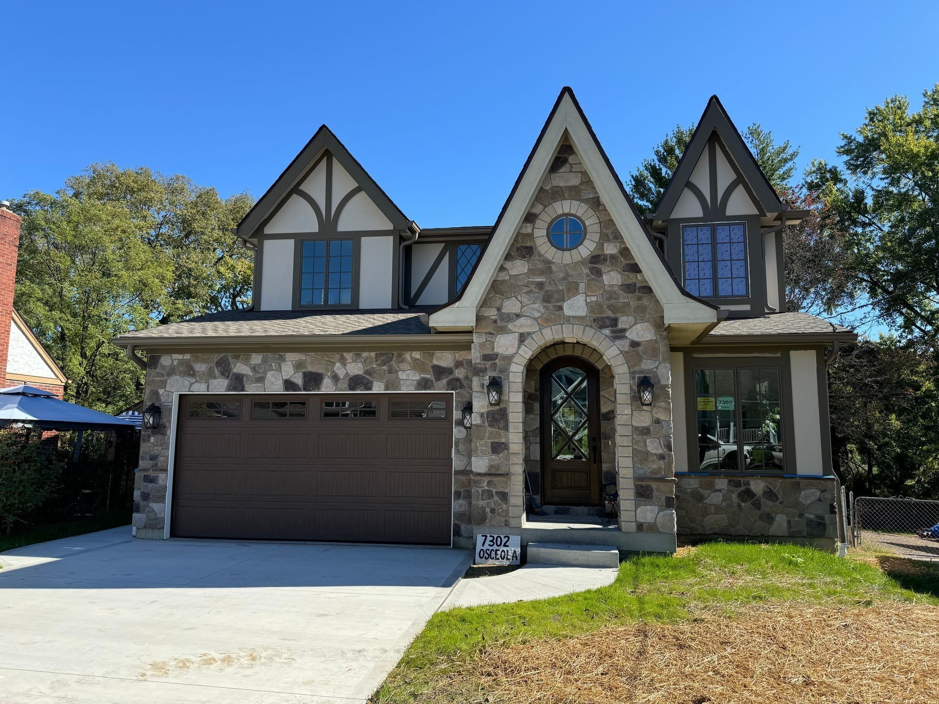 Stone and stucco Tudor-style house with arched doorway and attached garage, on a sunny day.