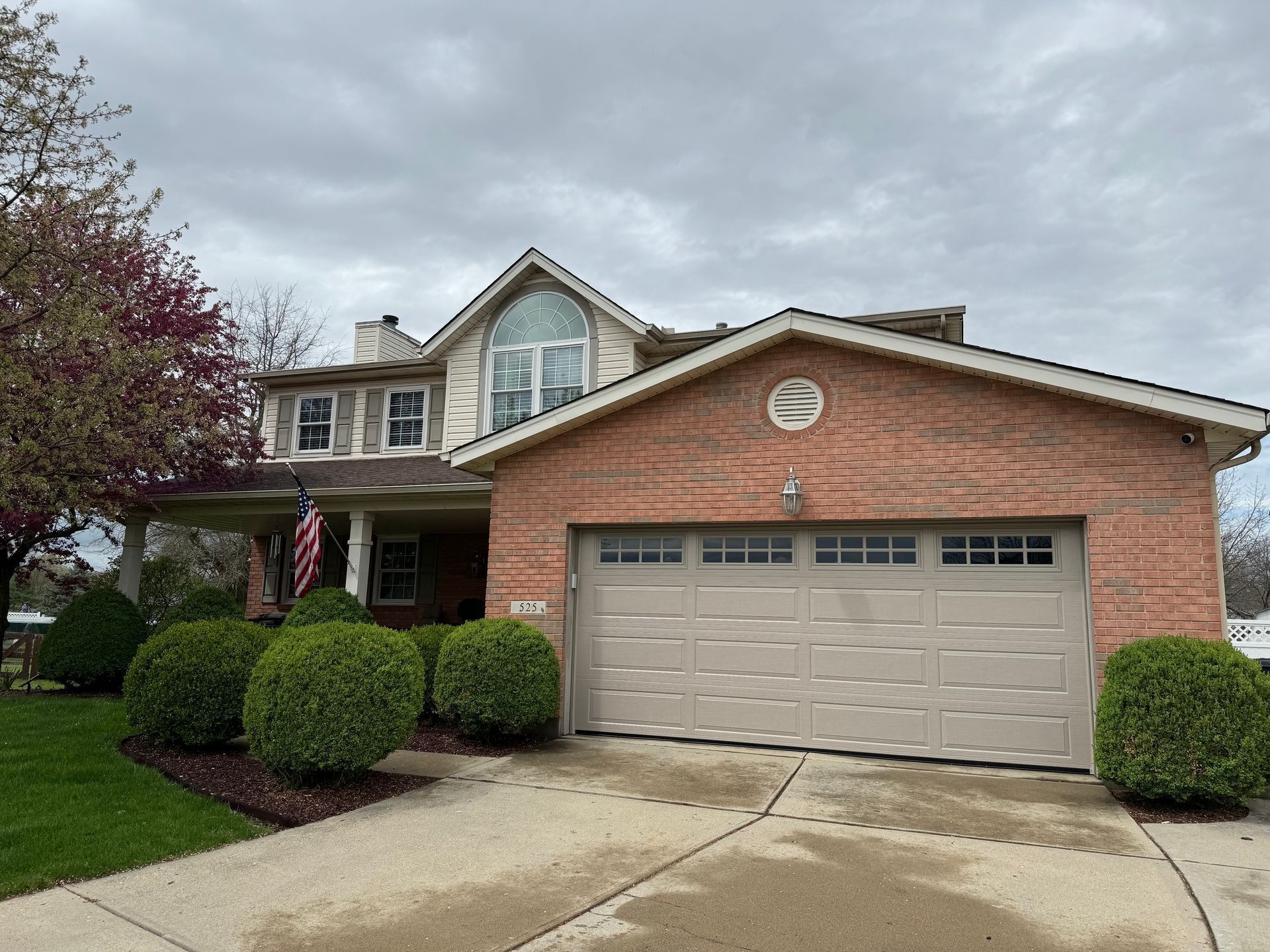 Two-story brick house with beige garage door, arched window, and landscaped yard under a cloudy sky.