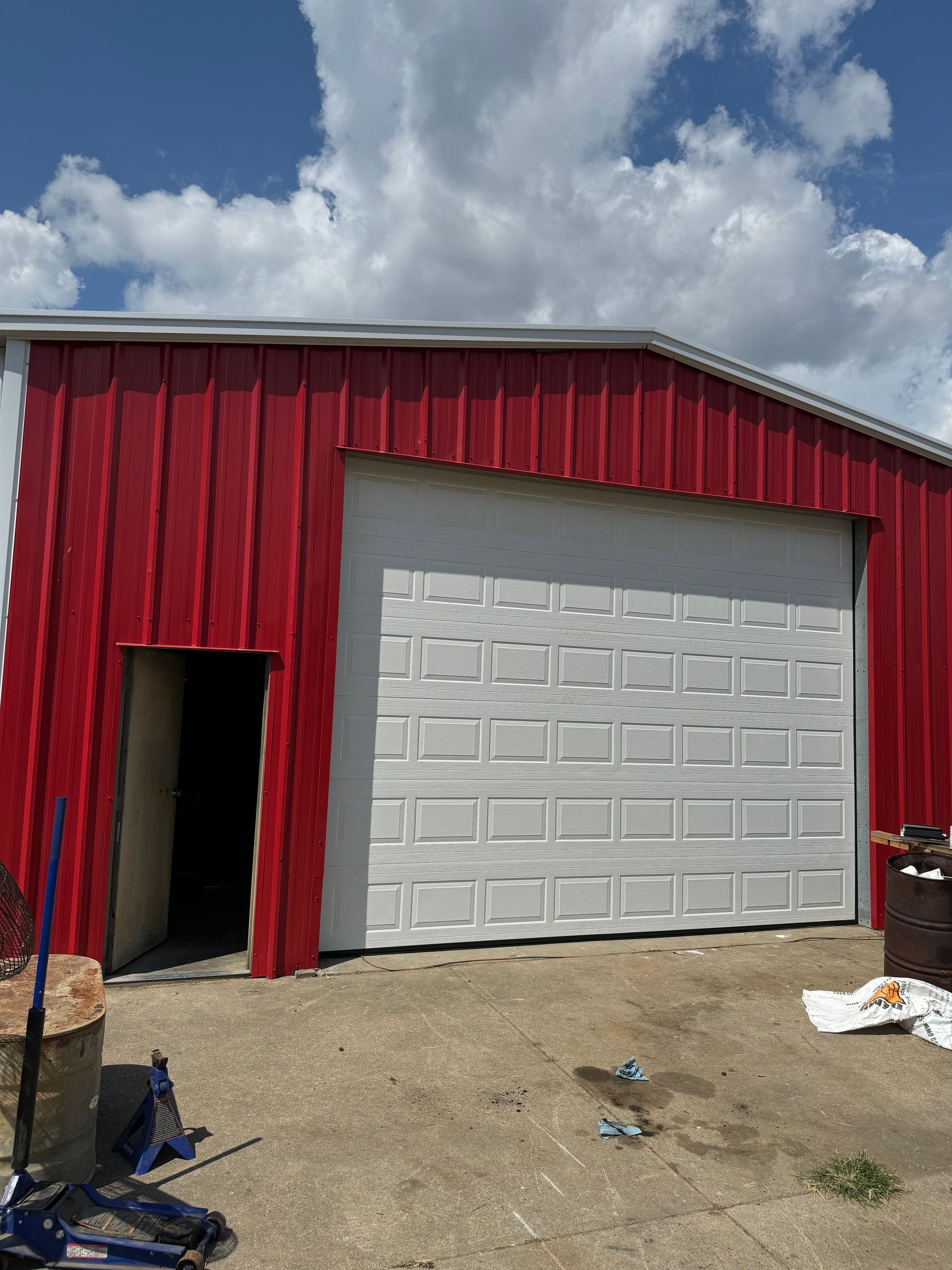 Red and white metal building with a garage door and an open door. Blue sky with clouds overhead.