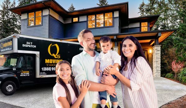 A family is standing in front of a house with a moving truck in the background.