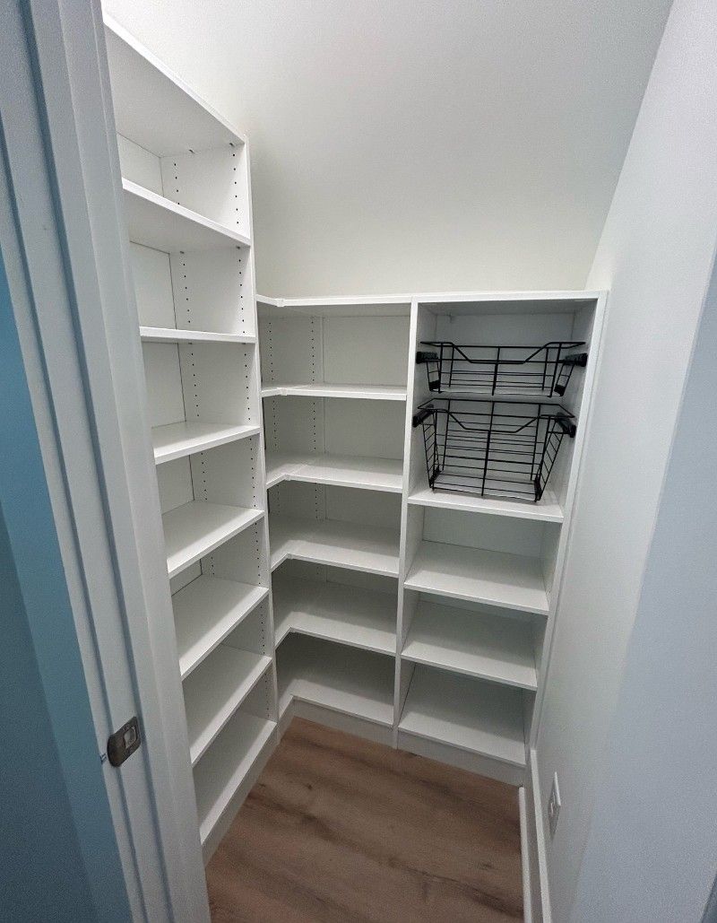 A small closet with white shelves and wooden floors under a staircase.