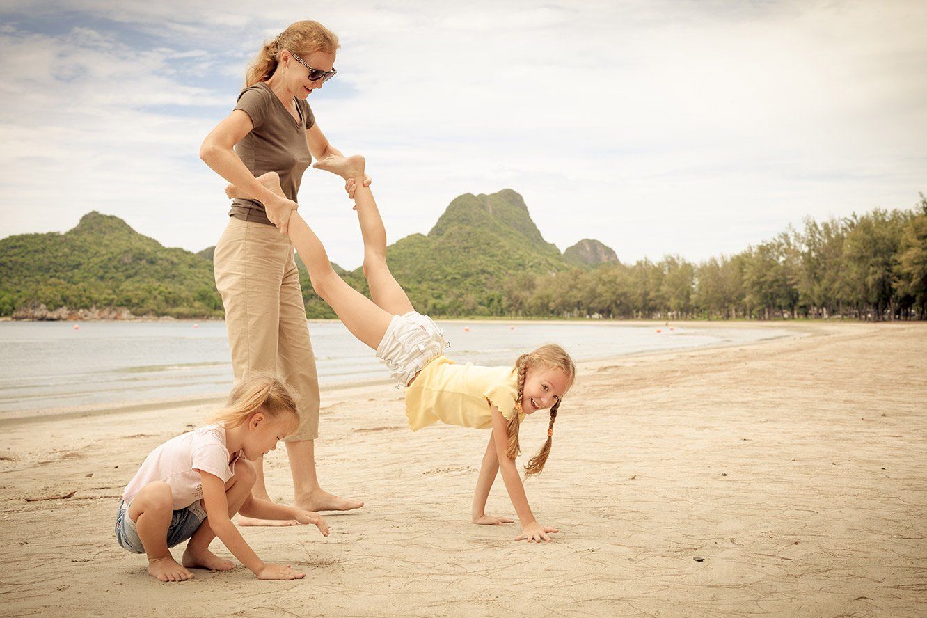 Woman helping a girl do a handstand on a beach, another girl playing in sand, mountains in background.