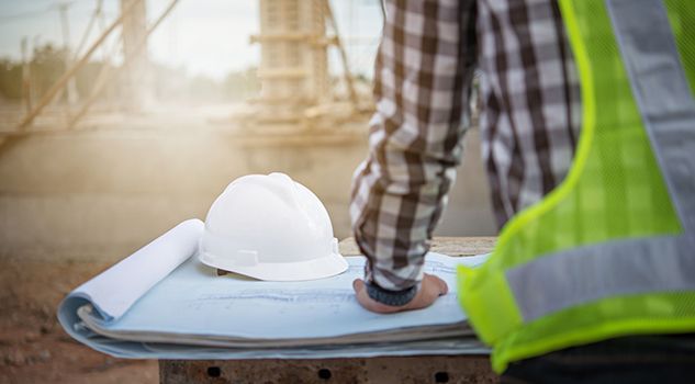 Construction worker in reflective vest reviewing blueprints beside a hard hat at a job site