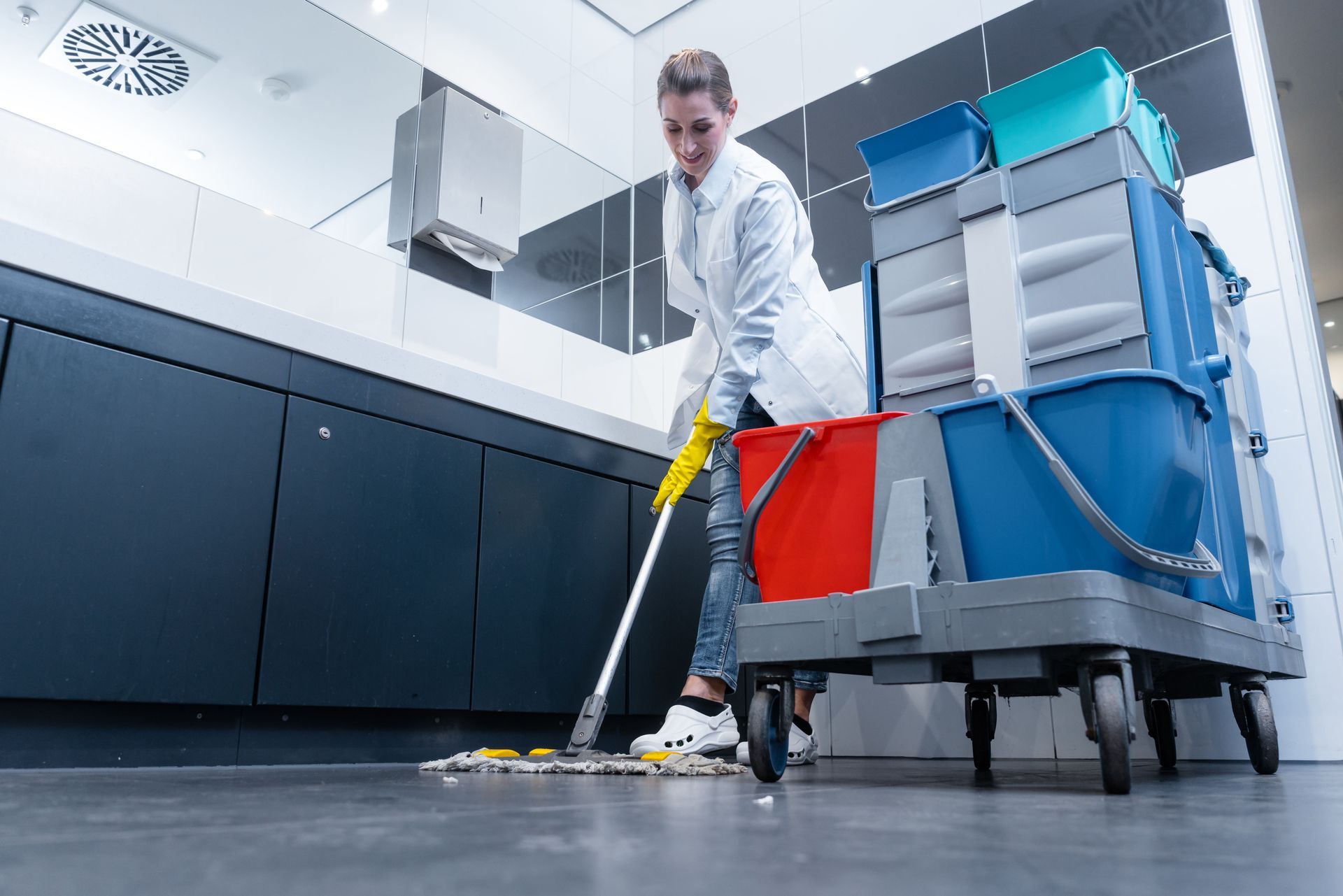 Woman mopping a bathroom floor, wearing gloves, and with a cleaning cart next to her.
