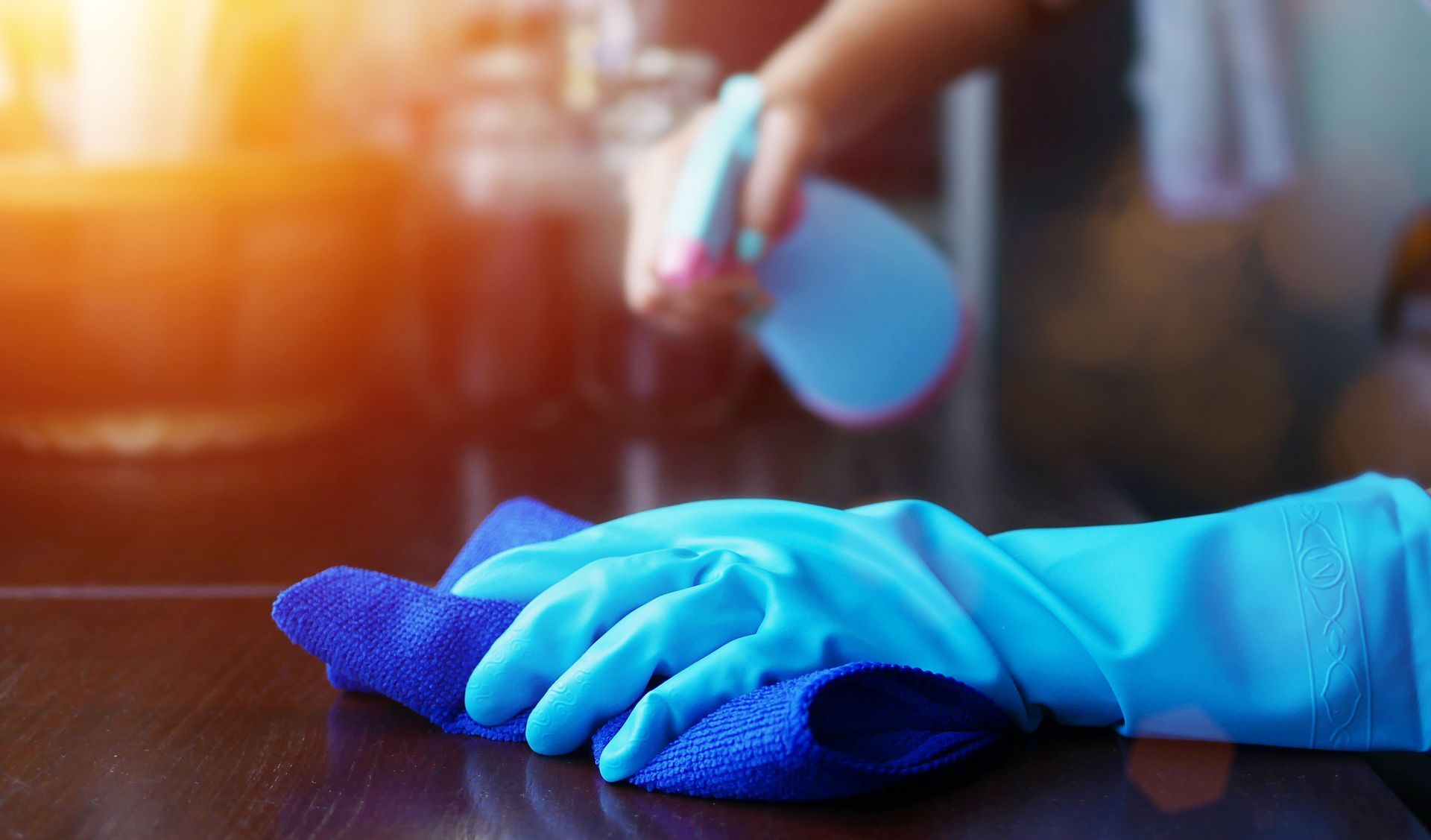 Person cleaning a wooden surface with a blue cloth and spray bottle, wearing blue gloves.