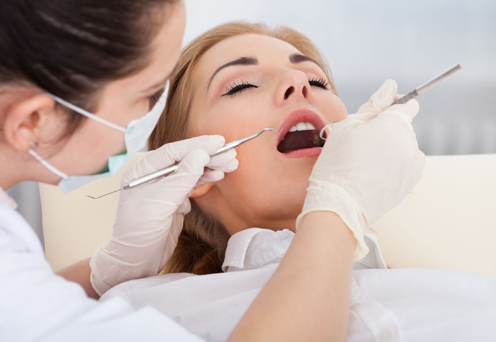 Female dentist performing a dental examination on a woman lying in a dental chair, using dental tools while wearing gloves and a face mask.