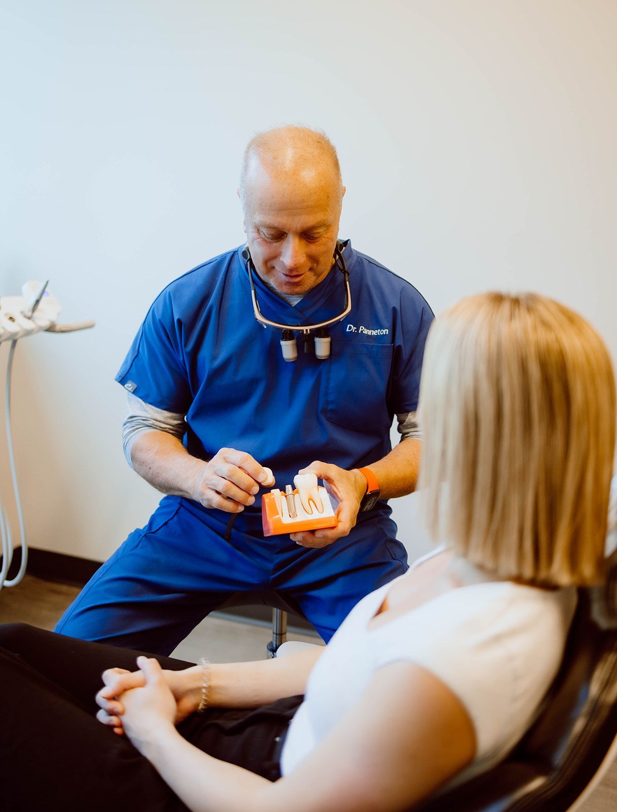 A dentist is talking to a woman in a dental chair while holding a model of teeth.