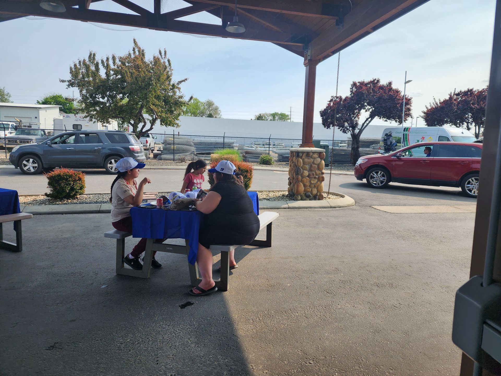People seated at a picnic table under a pavilion in a parking lot, with cars and trees in the background.