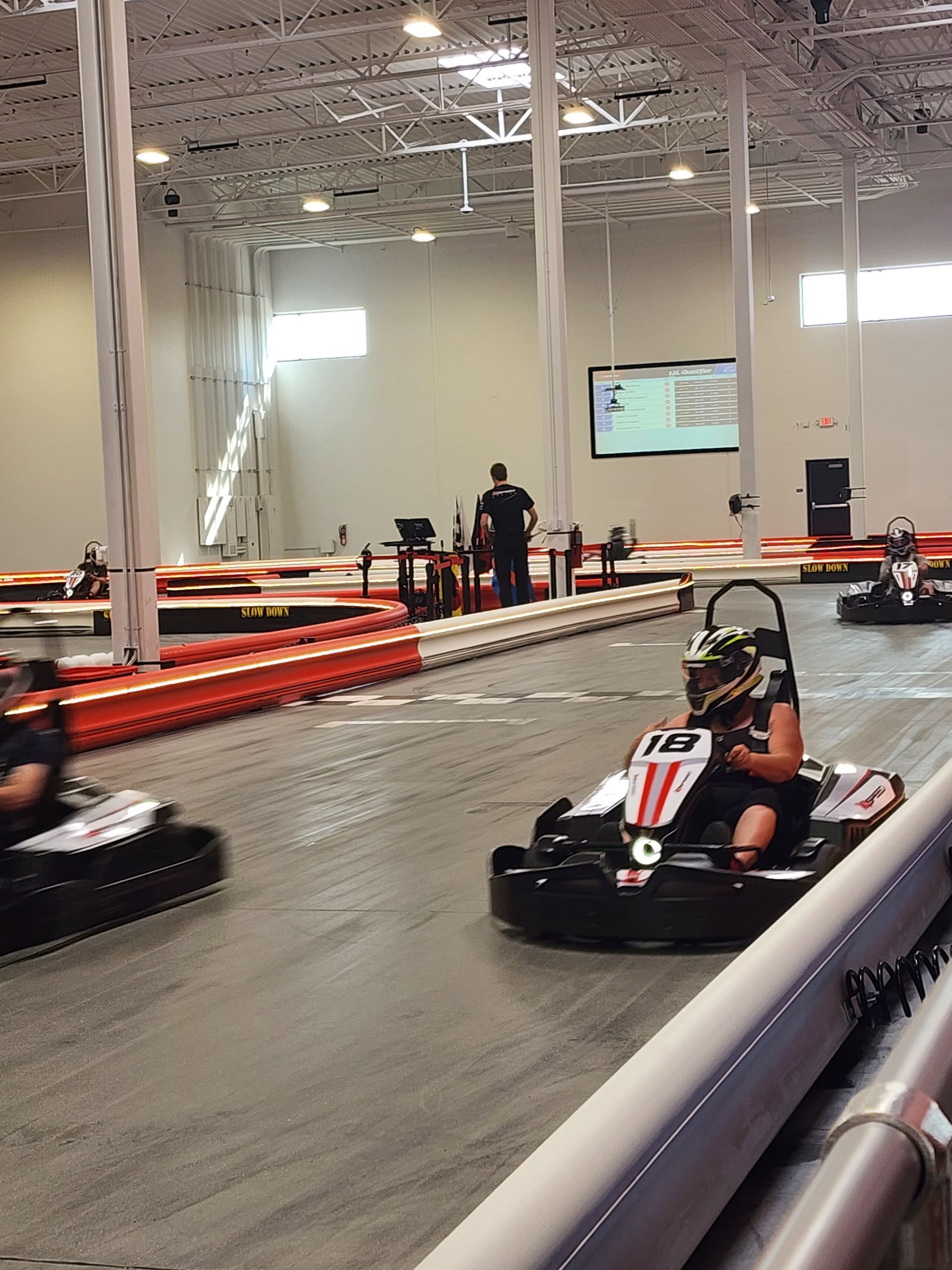 Go-karts racing on an indoor track, with a black kart in the foreground and orange barriers lining the course.