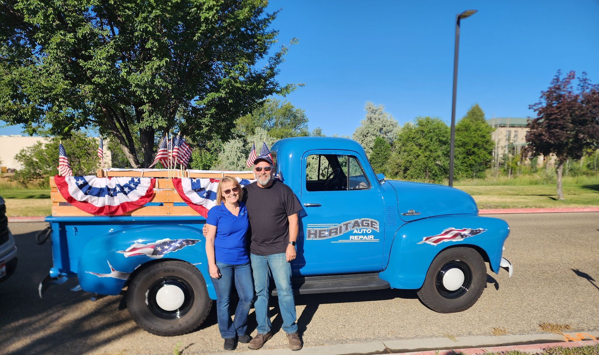 Two people pose beside a bright blue vintage pickup decorated with red, white, and blue bunting outdoors.