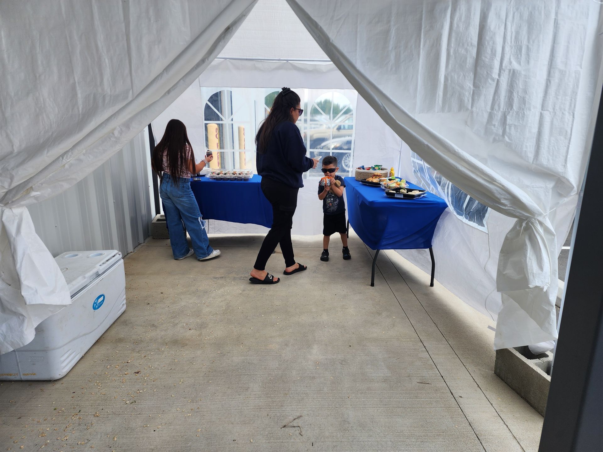 People browsing tables under a white canopy tent with blue tablecloths.