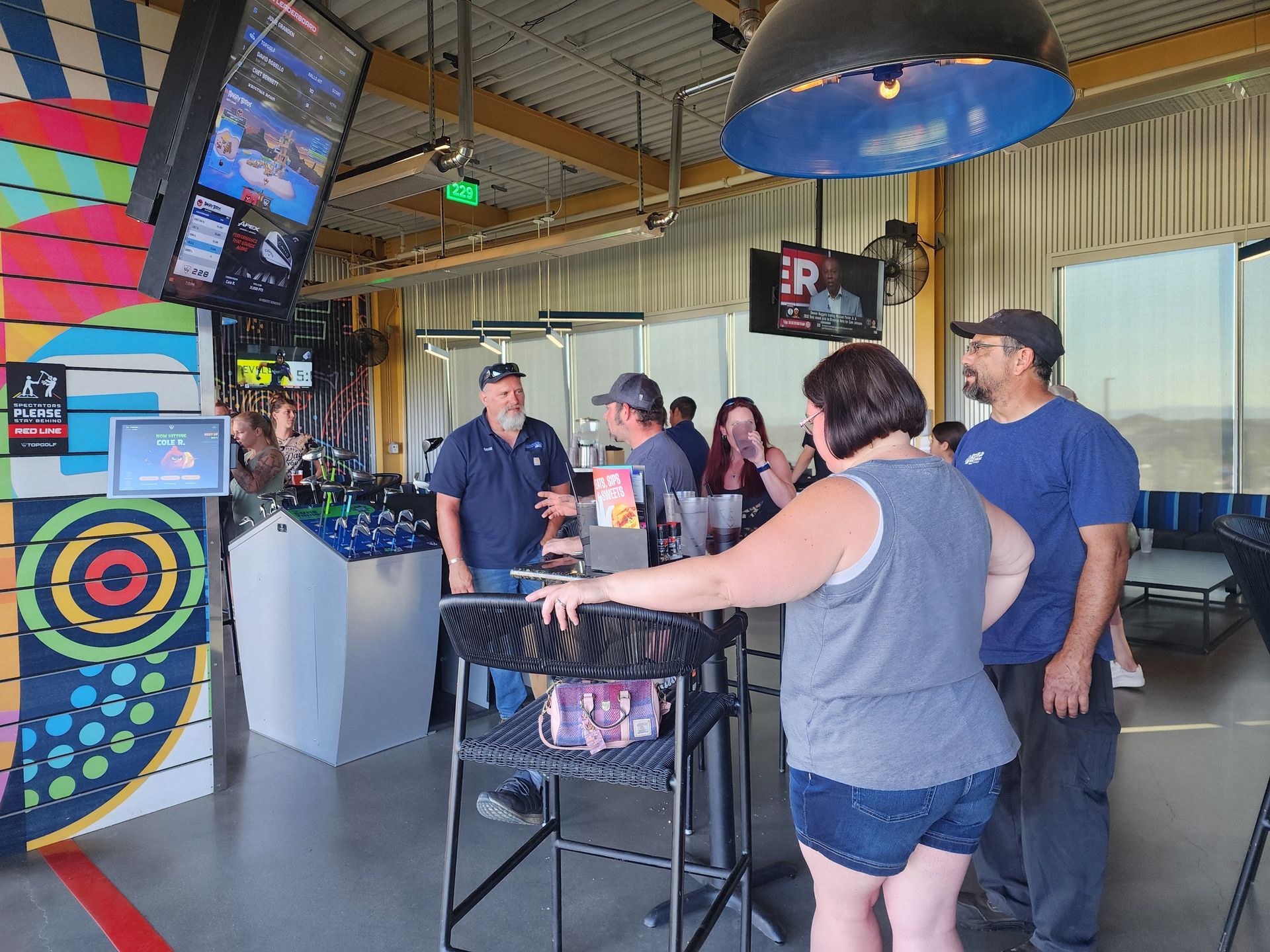 People socializing at a bar with dartboards and TVs, standing around a high table indoors.