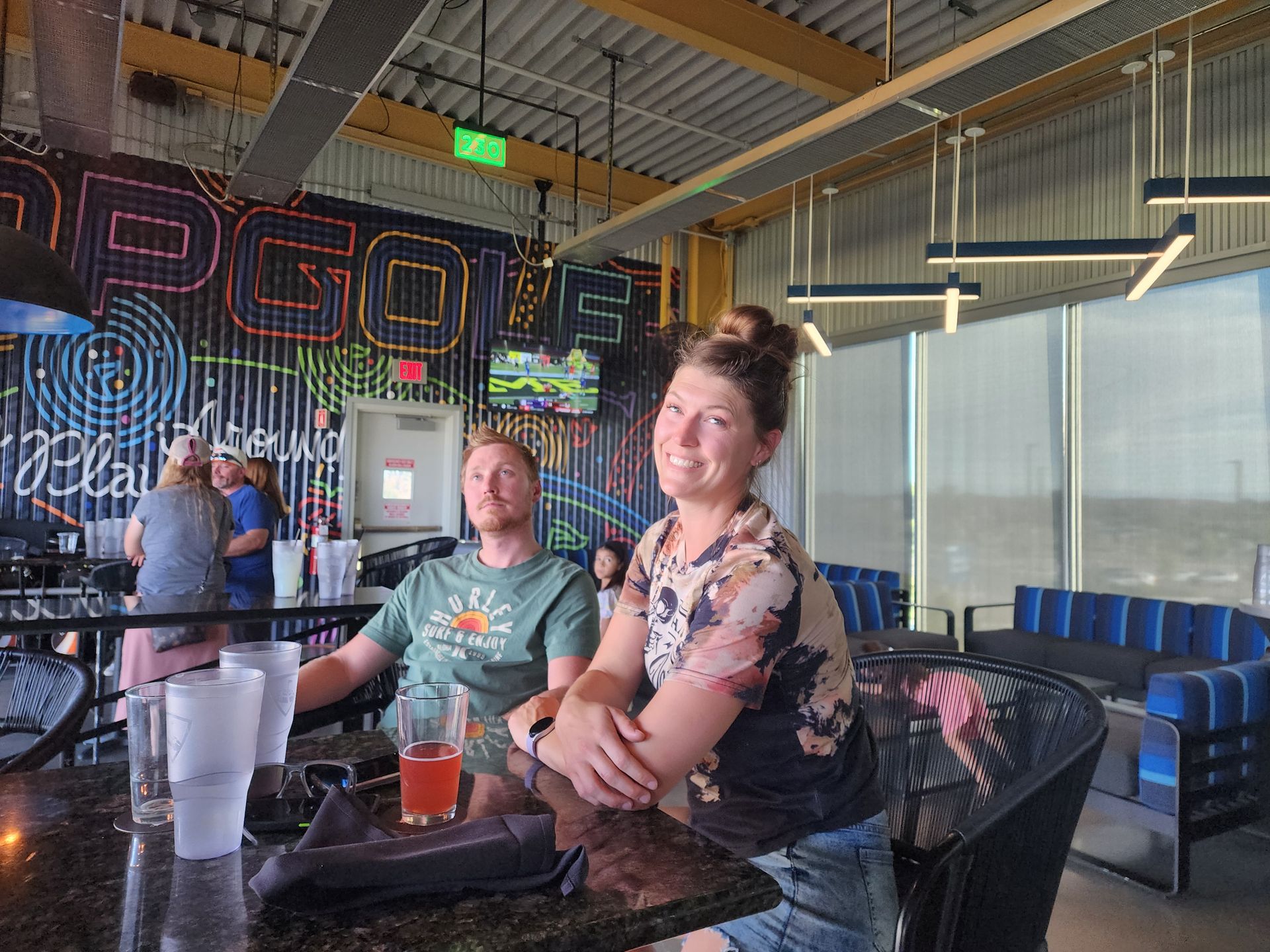 Two people seated at a bar table in a neon-lit indoor venue, smiling with drinks nearby.