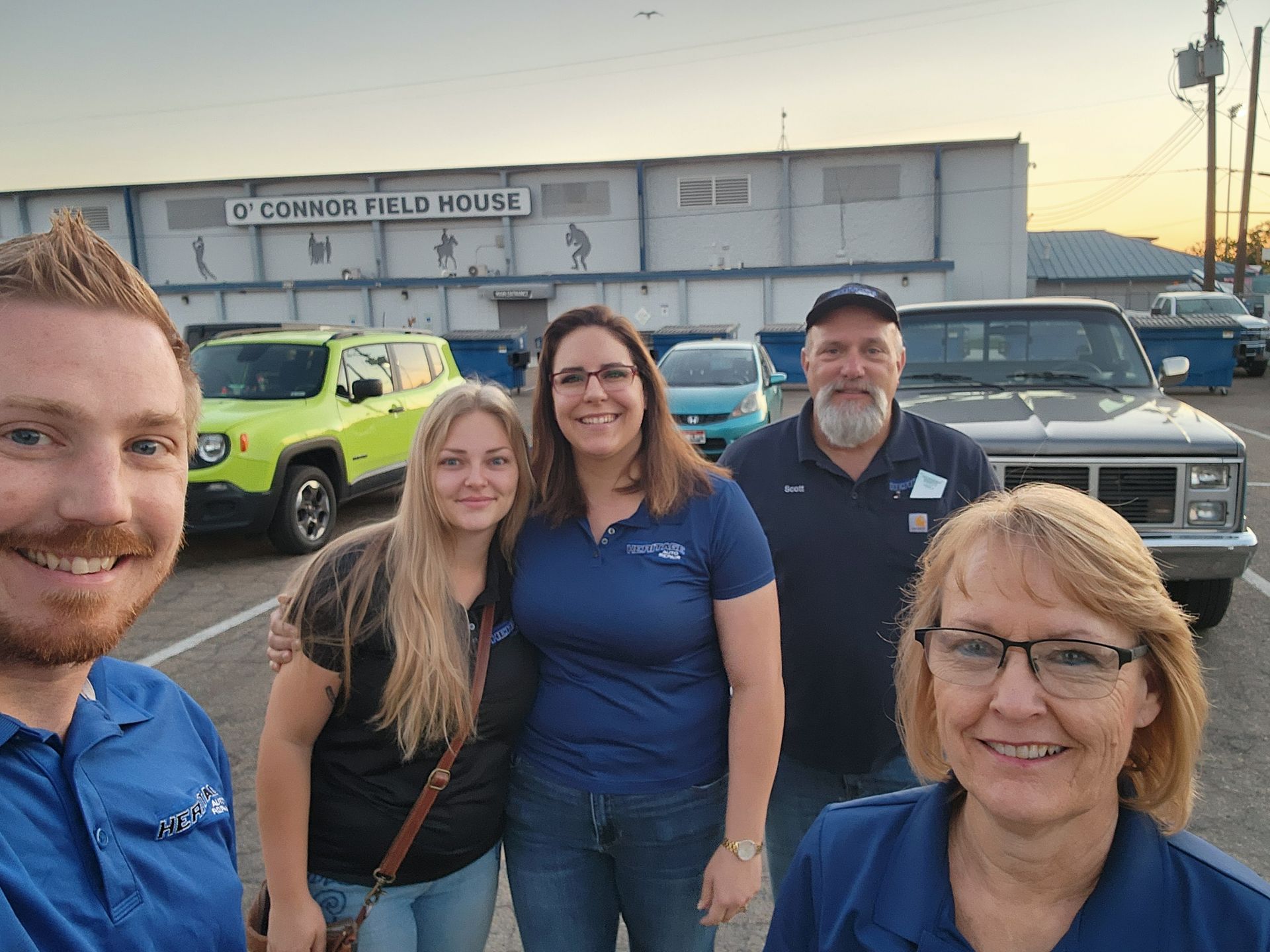 Five people posing in front of a field house and parked vehicles at sunset.