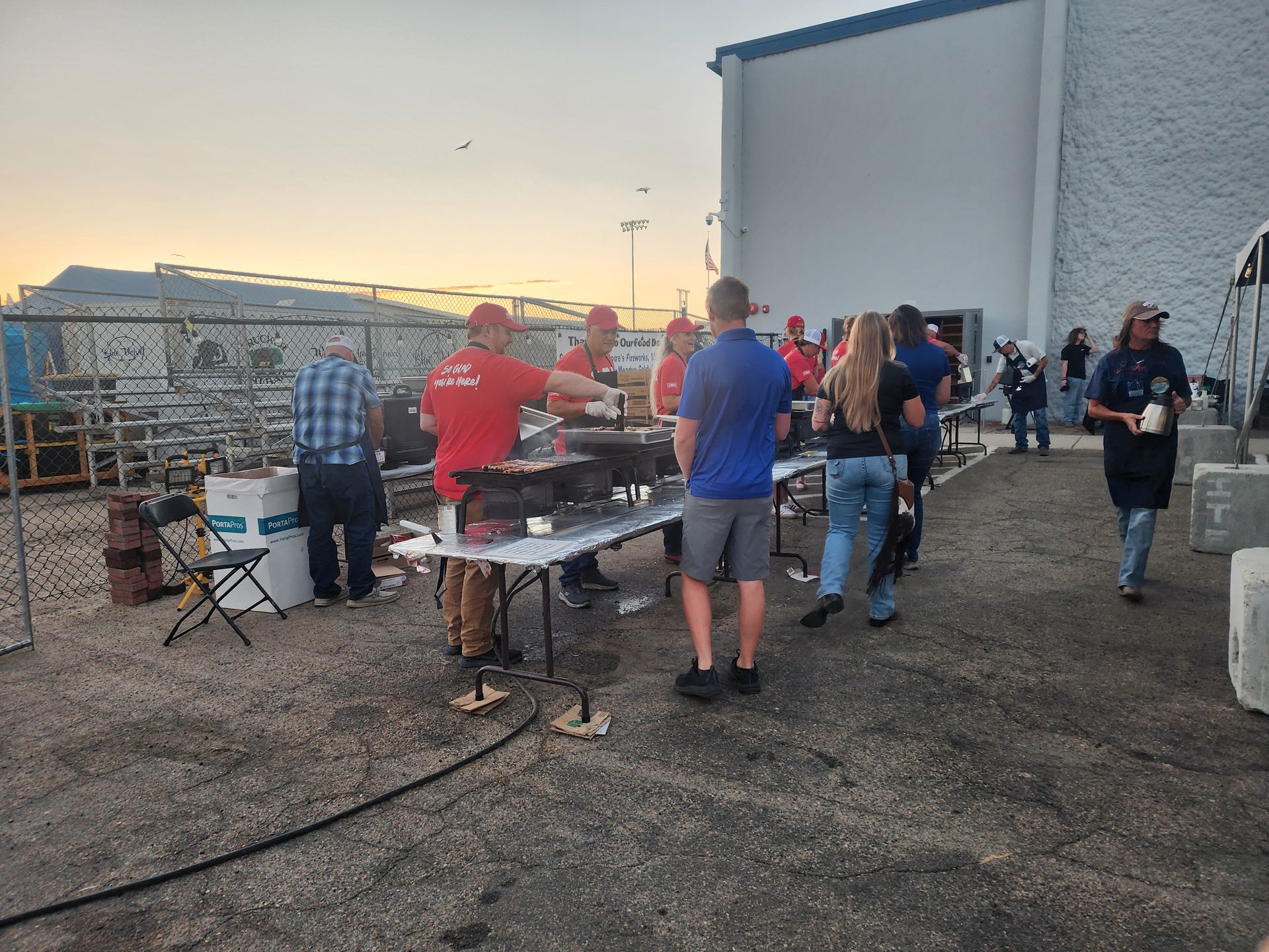 People grilling and chatting in a parking lot beside a white building at sunset.