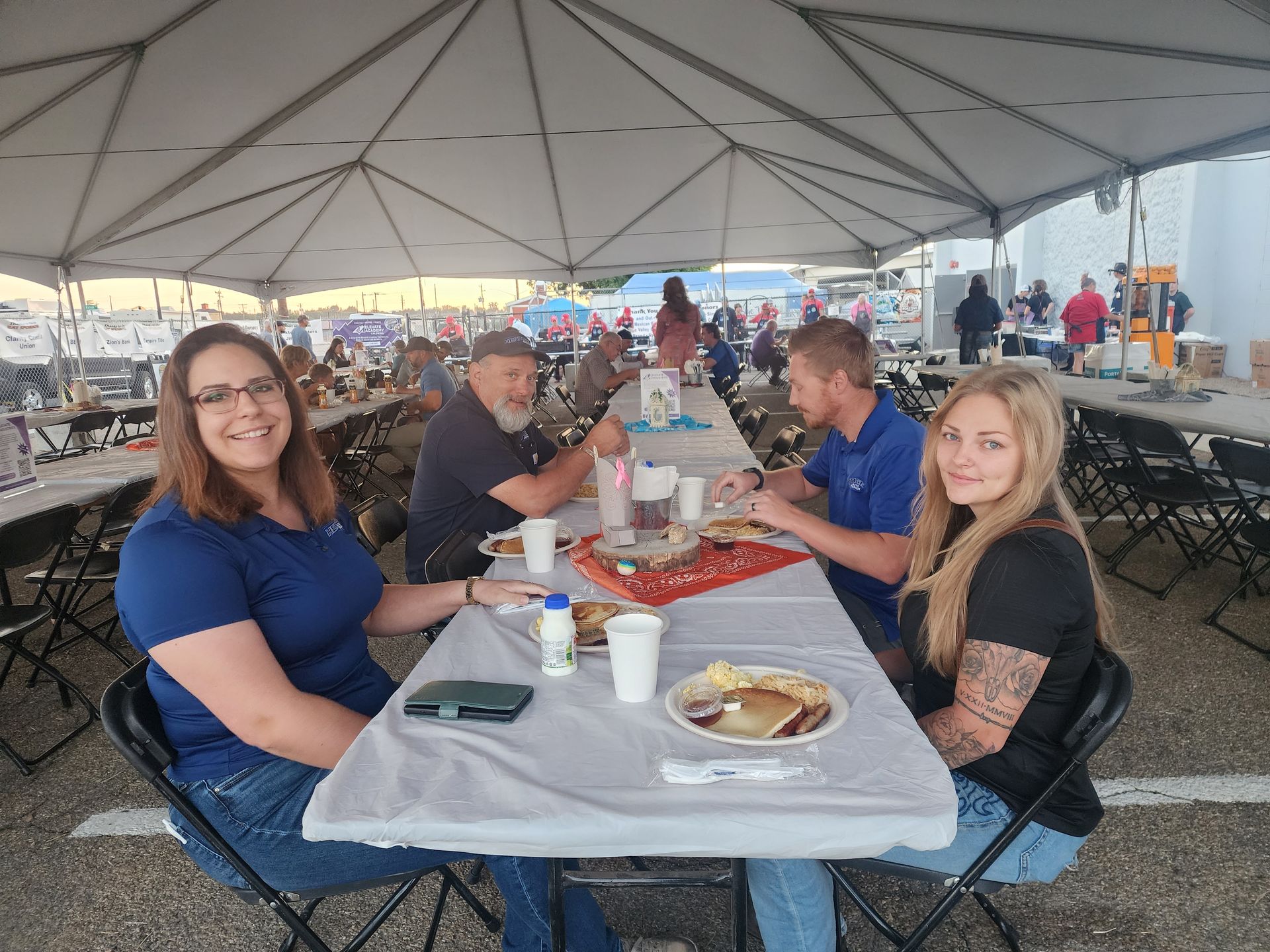 People seated at a picnic table under a large white event tent, eating and talking.