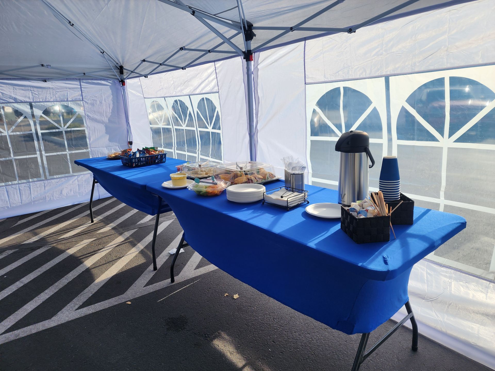 Buffet table with blue tablecloth under a white canopy, set with food trays, coffee urn, and cups.
