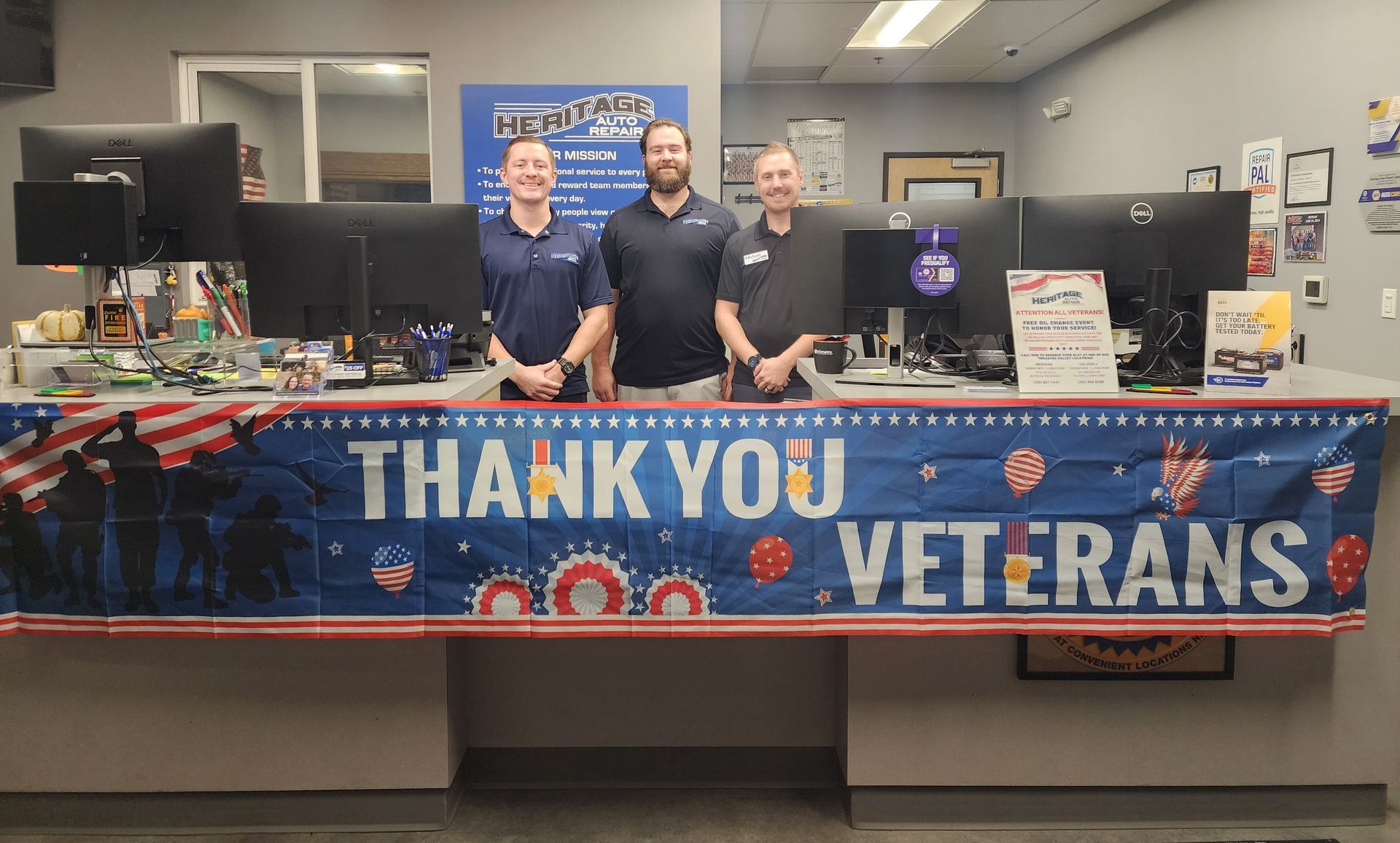 Two men behind a booth with a blue “THANK YOU VETERANS” banner and two computer monitors.