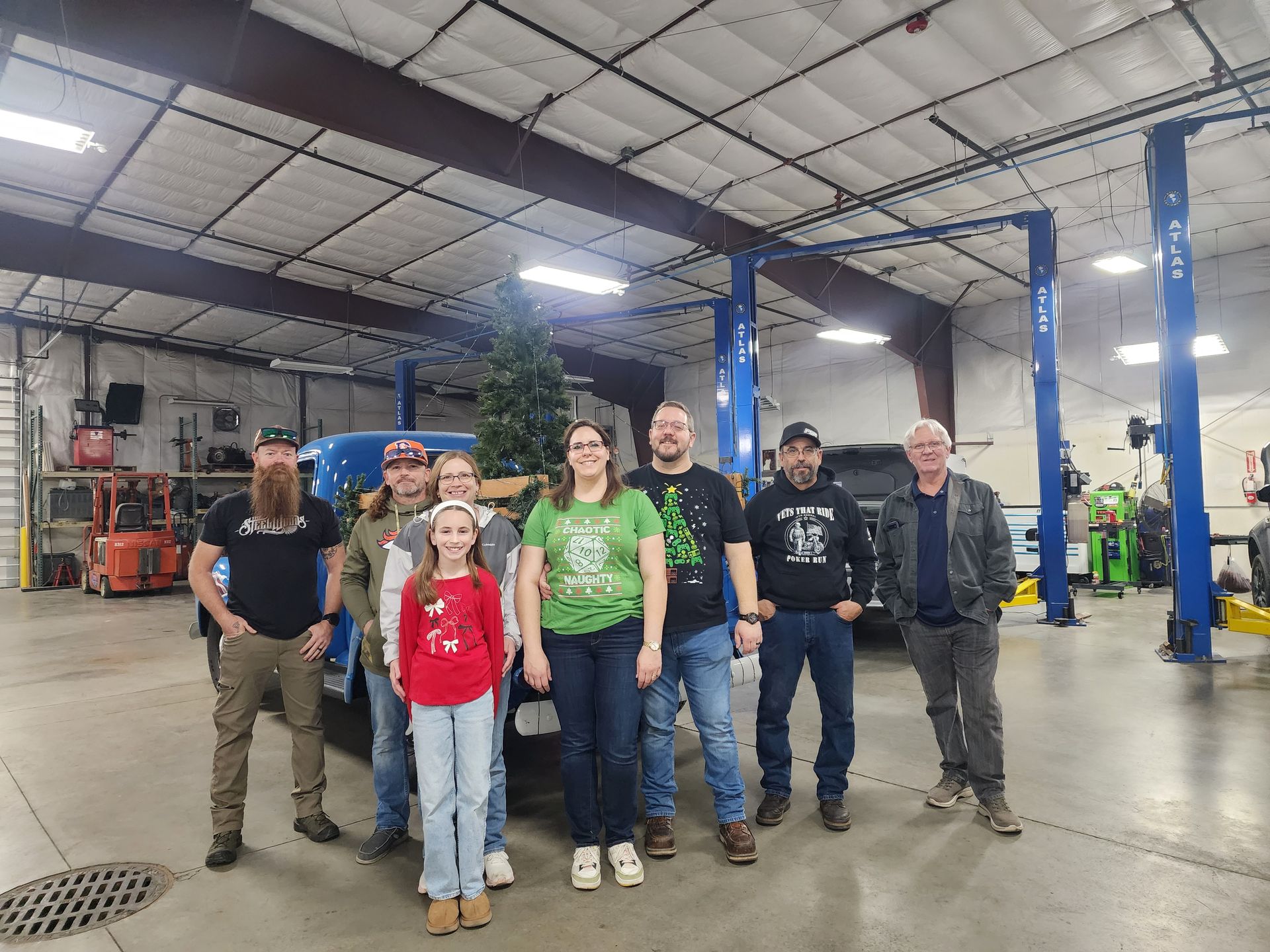 Group of six people standing in an auto repair shop with blue vehicle lifts and a car in the background.