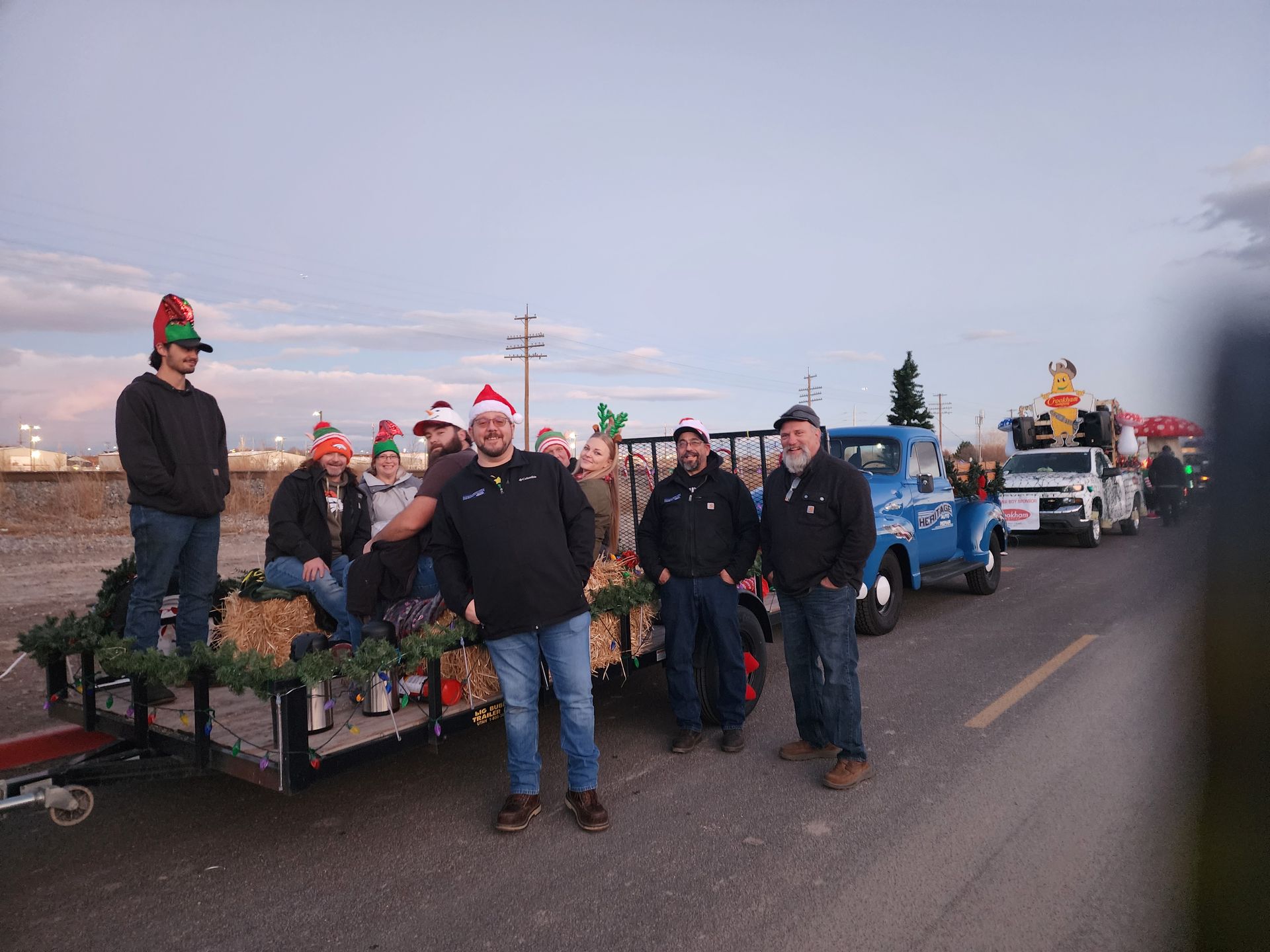 People in Santa hats standing by decorated trailers and trucks on a roadside at dusk.