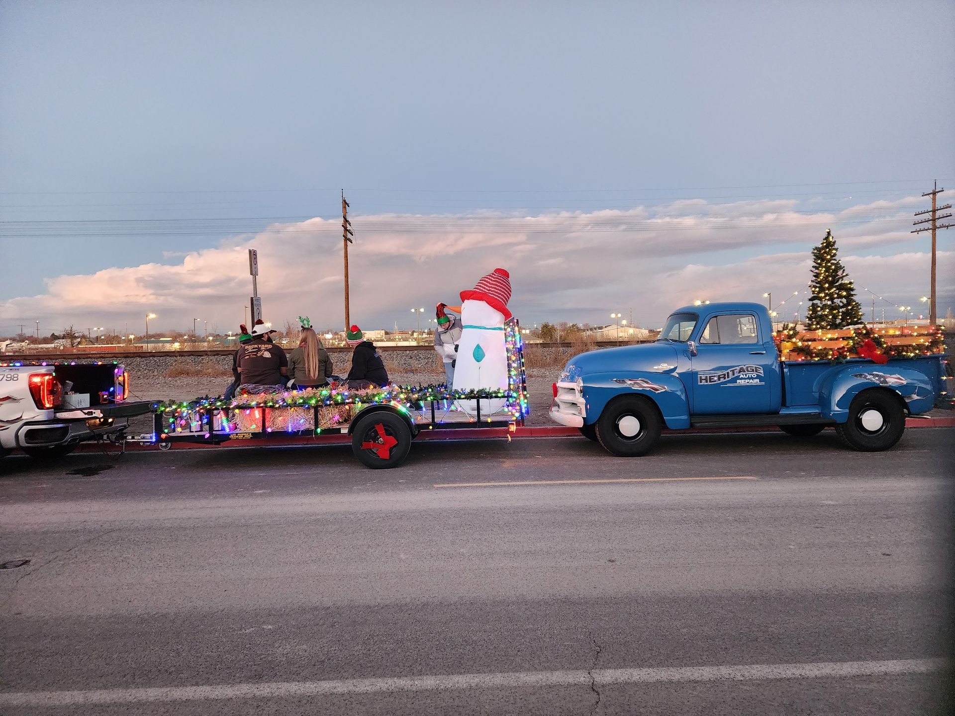 Blue pickup towing Christmas parade float with Santa and decorated trees along a road at dusk.