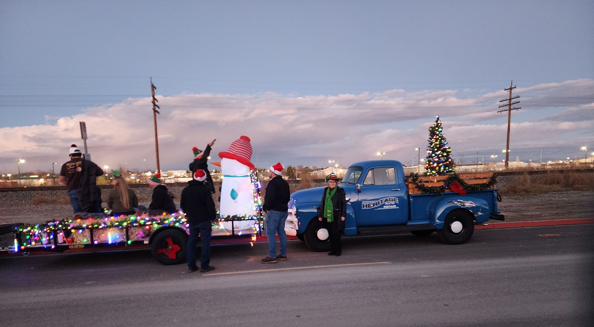 Decorated blue pickup truck in a holiday parade with people standing beside it at dusk.