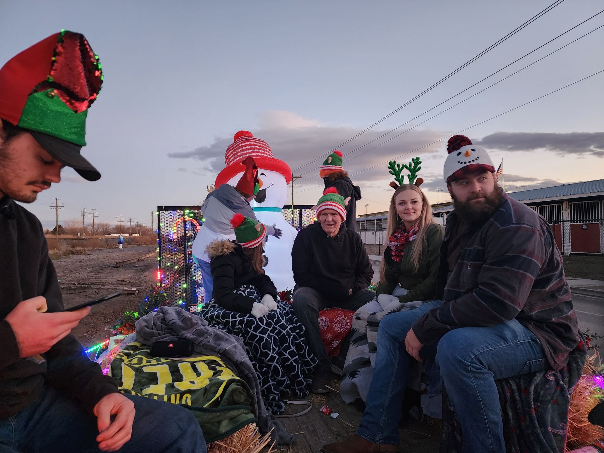 Group of people in festive holiday hats sitting outdoors at dusk with lights and decorations.