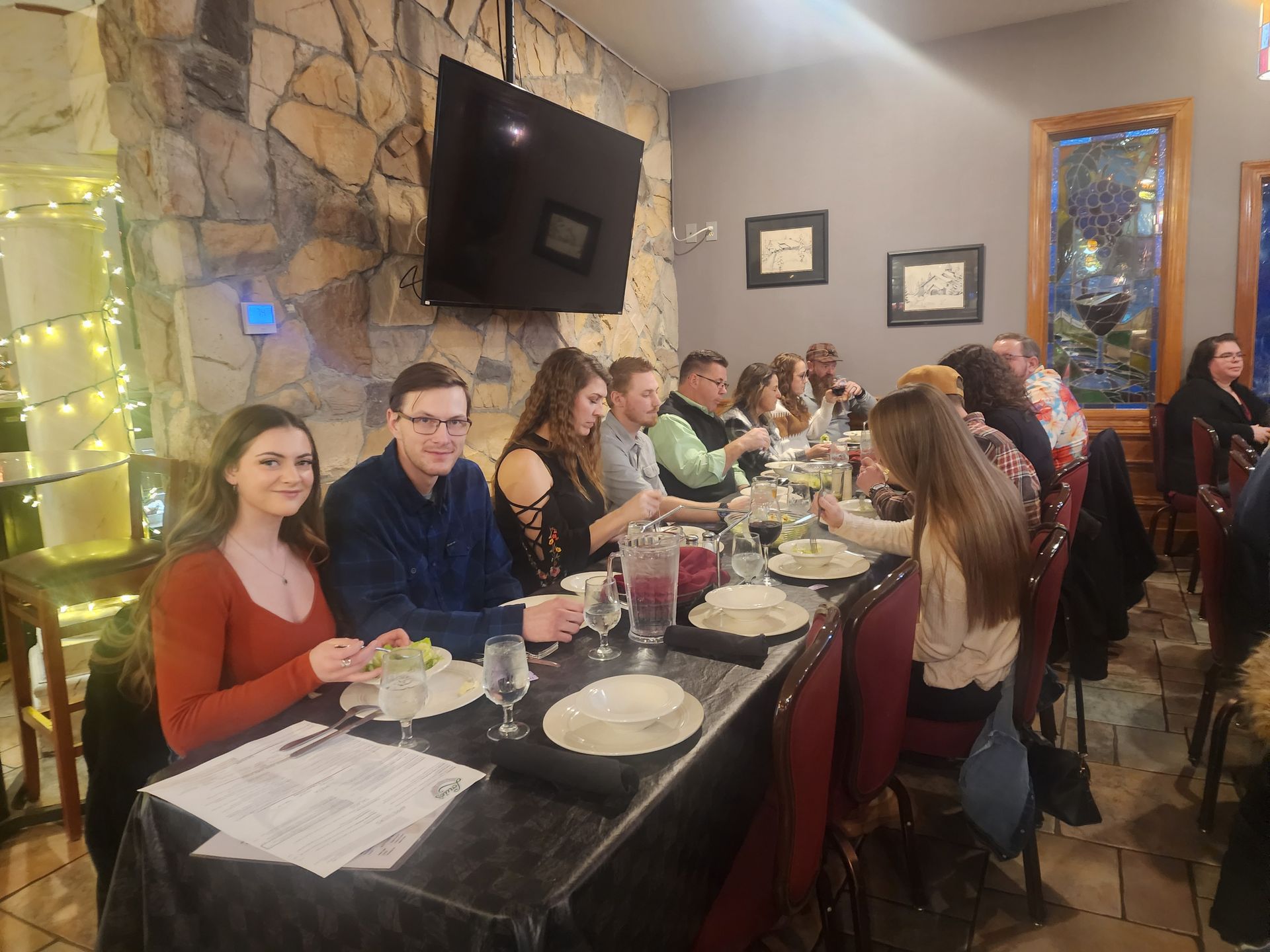 Restaurant table with diners seated along a long black-clothed table beside a stone wall and TV.