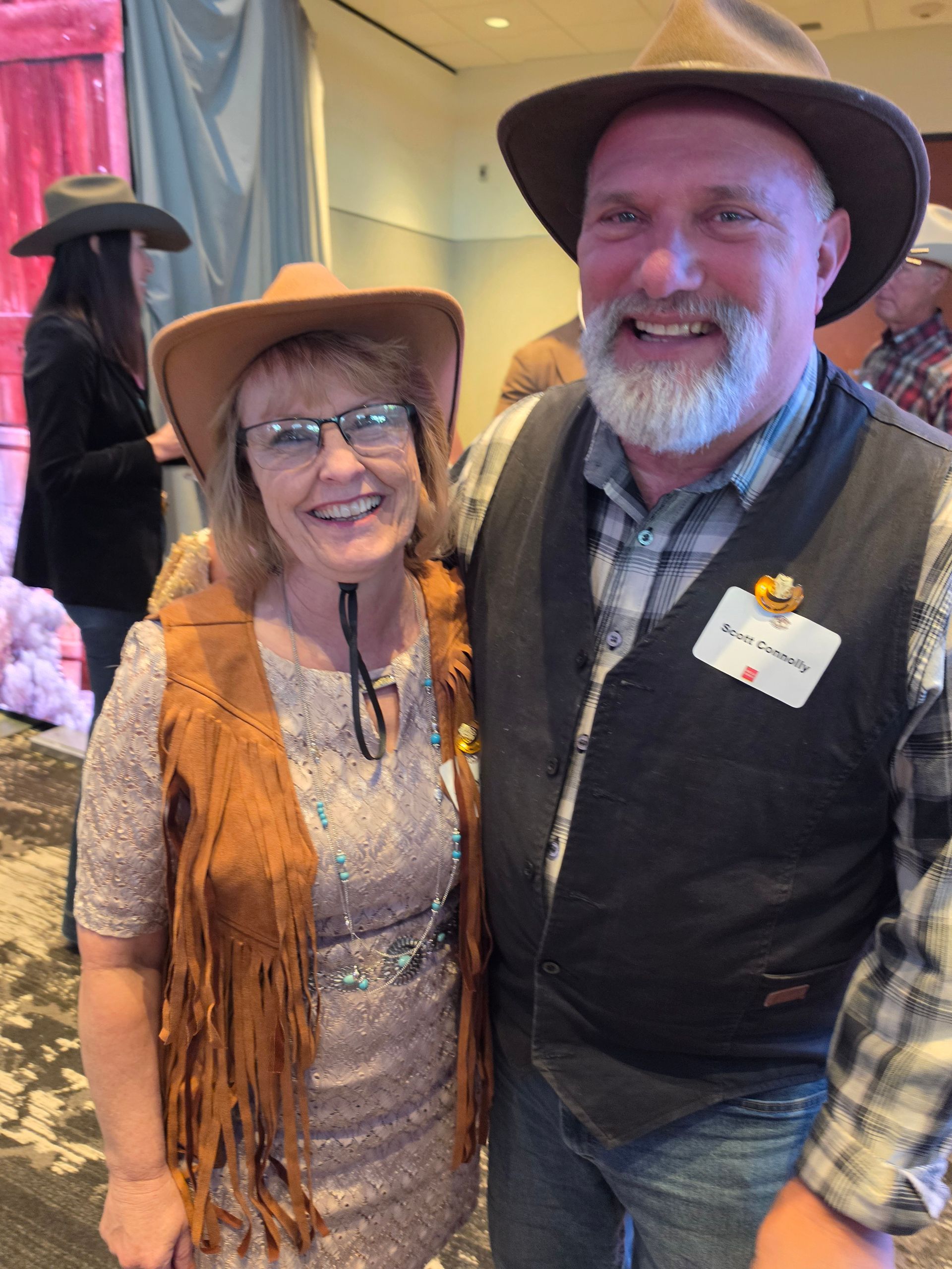 Two smiling people in cowboy hats stand together indoors at a western-themed event.