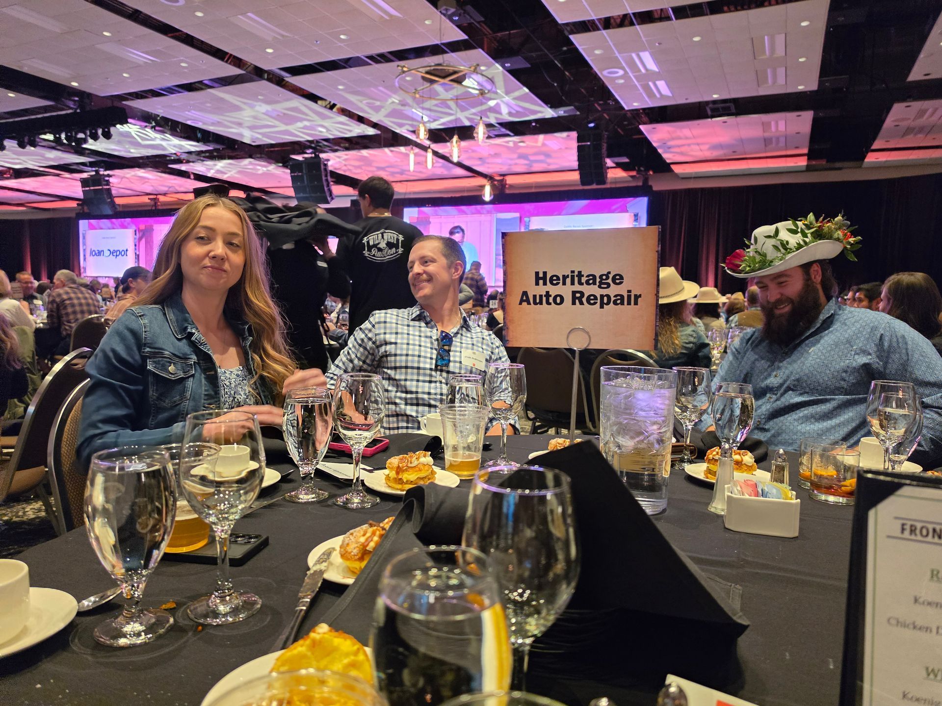 Three people seated at a banquet table at an auto repair event with a Heritage Auto Repair sign.