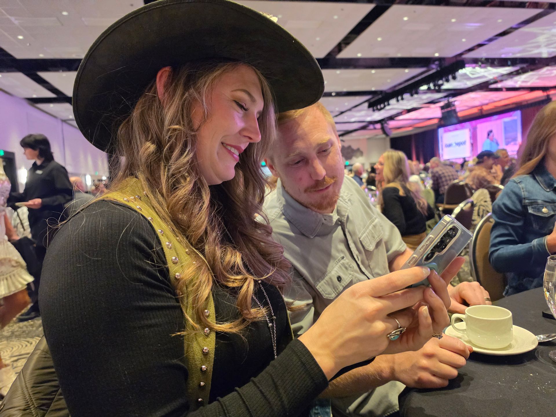 Two people smiling at a table, looking at a phone in a lively indoor event hall.