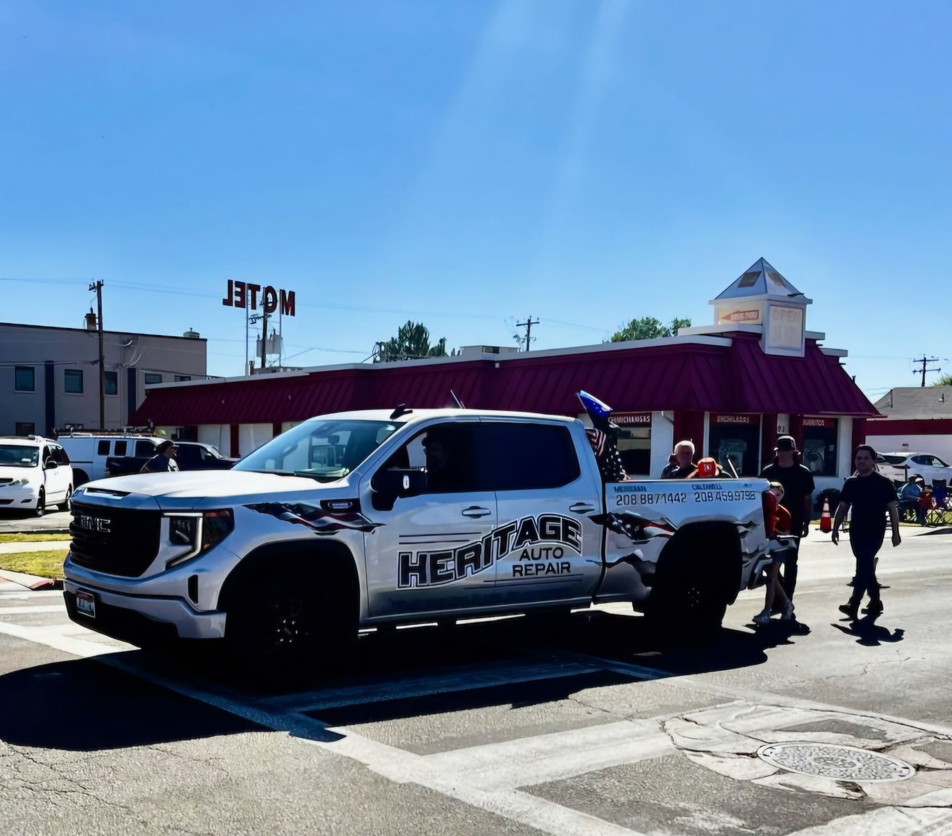 White pickup truck with Heritage logo at an outdoor event, with people walking nearby under a sunny sky.
