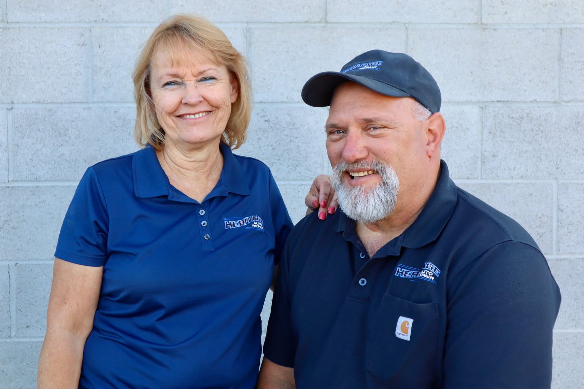 Two people in navy shirts smiling against a light gray brick wall.
