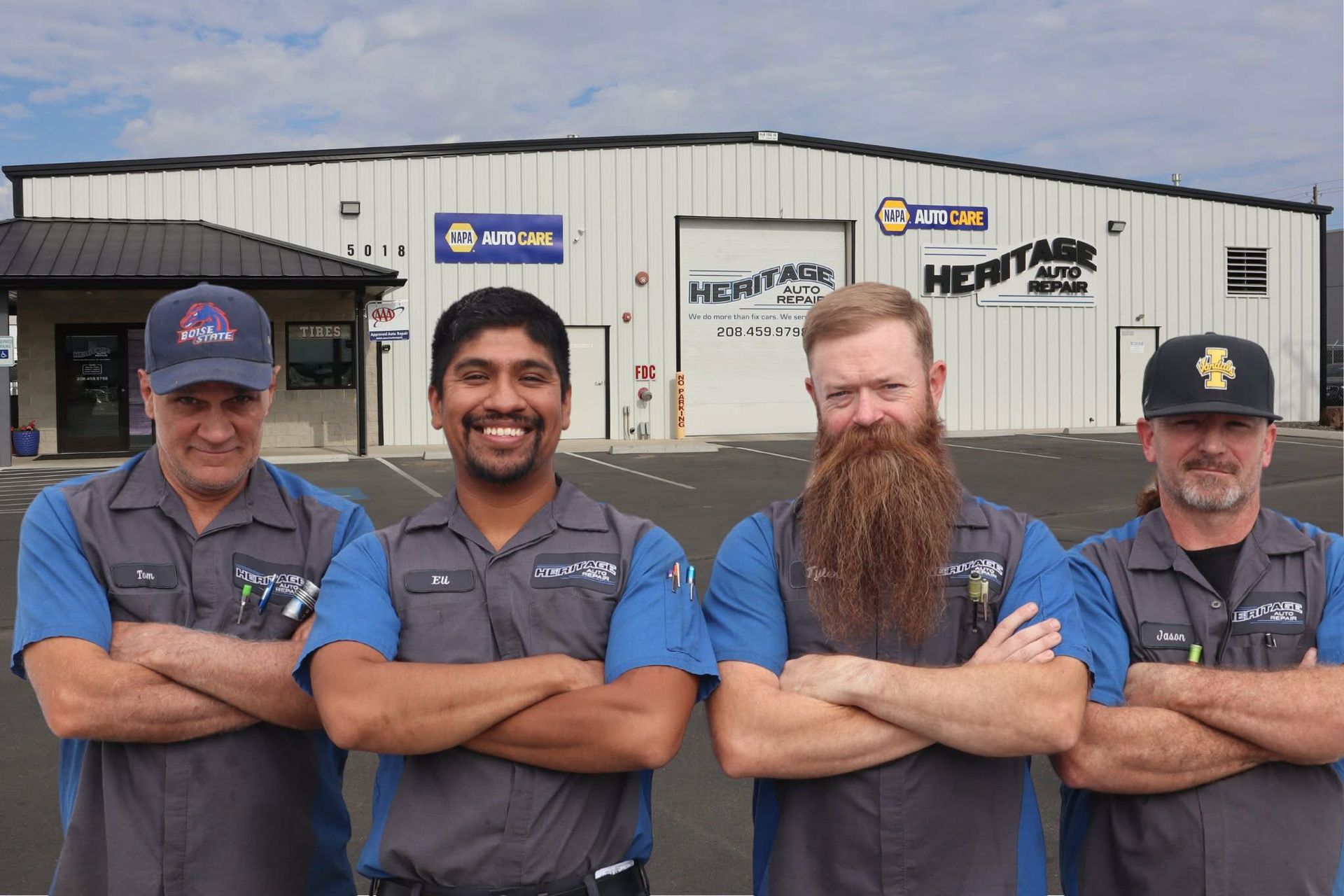 Four mechanics in gray and blue uniforms stand arms crossed outside a repair shop.