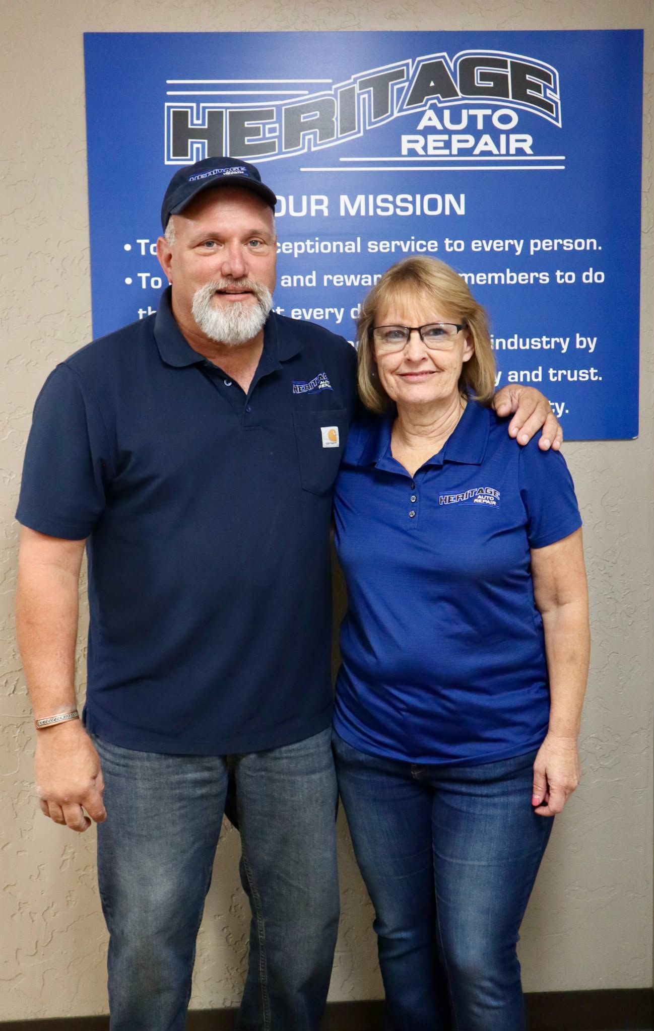 Two people standing in front of an auto repair shop sign, both wearing navy polo shirts and jeans.