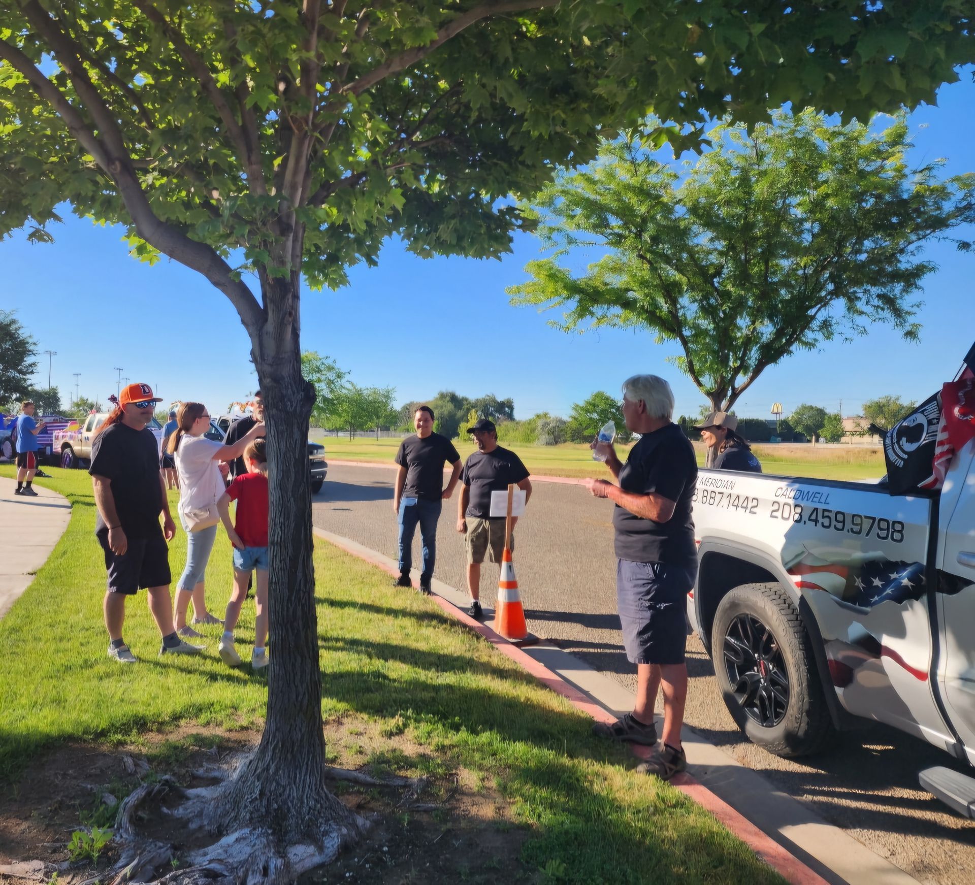 People standing by a white pickup truck on a sunny roadside with trees and orange cones nearby.